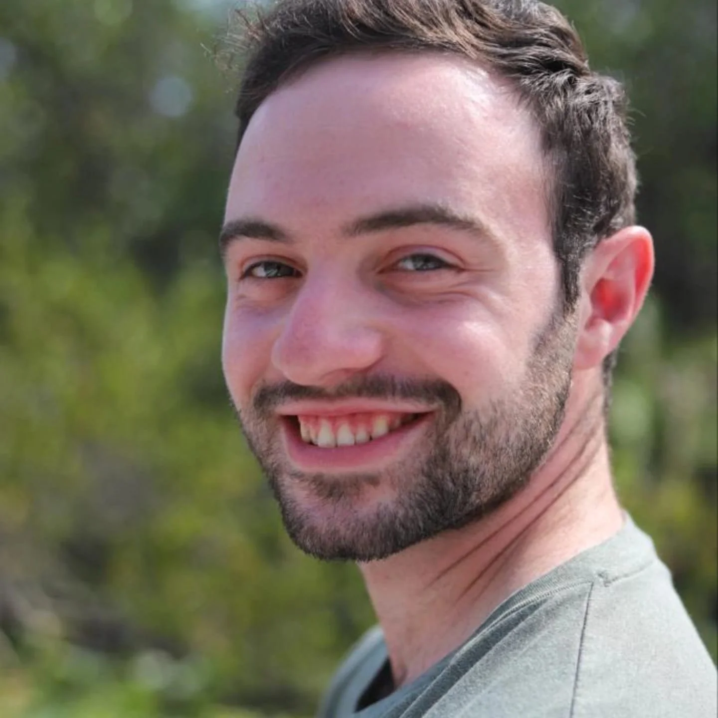 Close-up of a smiling man with short dark hair and a beard, outdoors with greenery in the background.