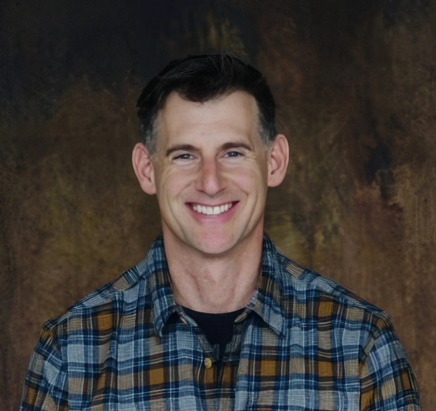 A smiling man with dark hair, wearing a plaid shirt, standing in front of a textured brown background.
