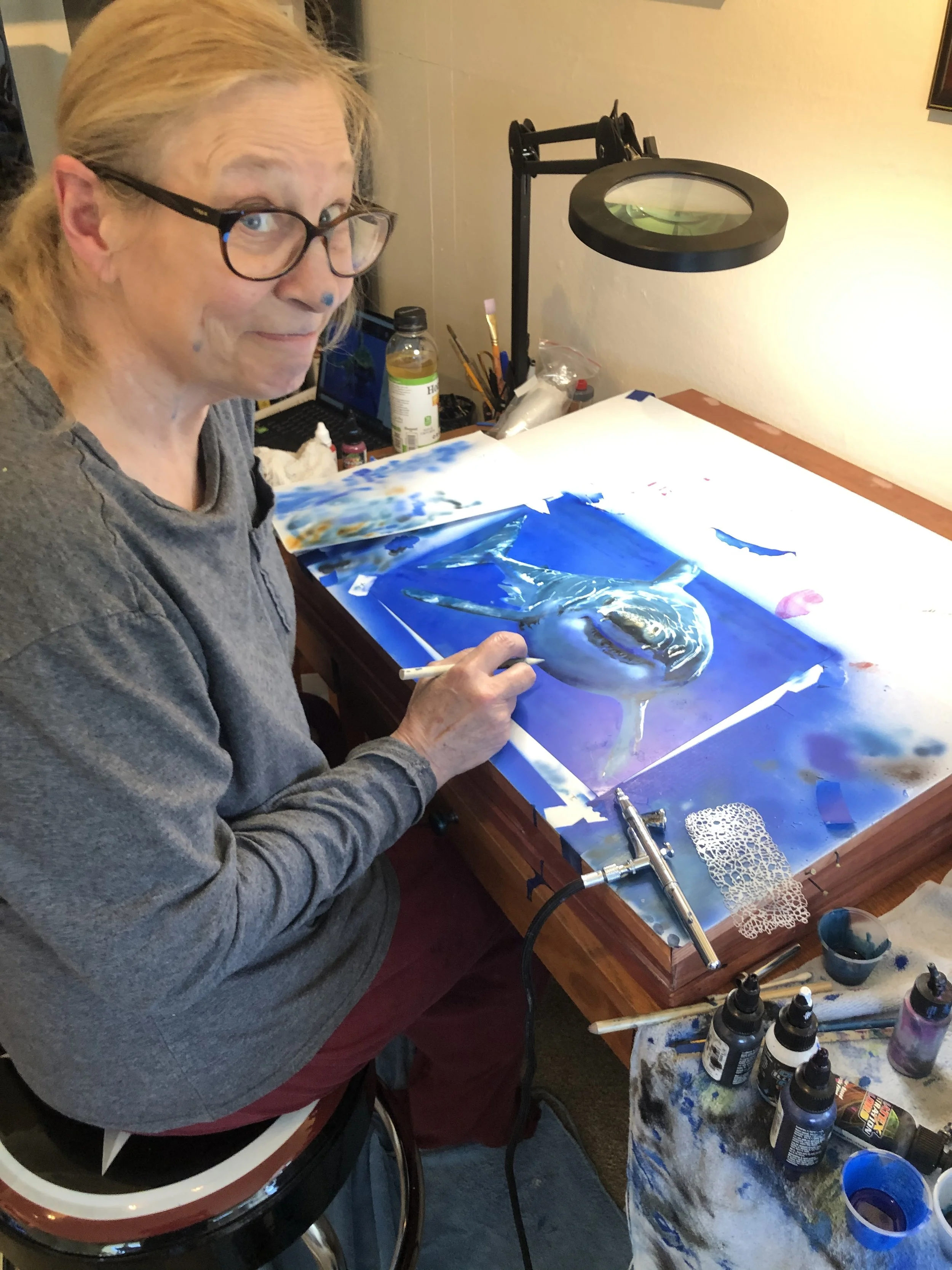 A woman with glasses sitting at a table creating a detailed painting of a dolphin on a lightbox. Art supplies and a lamp are on the table.