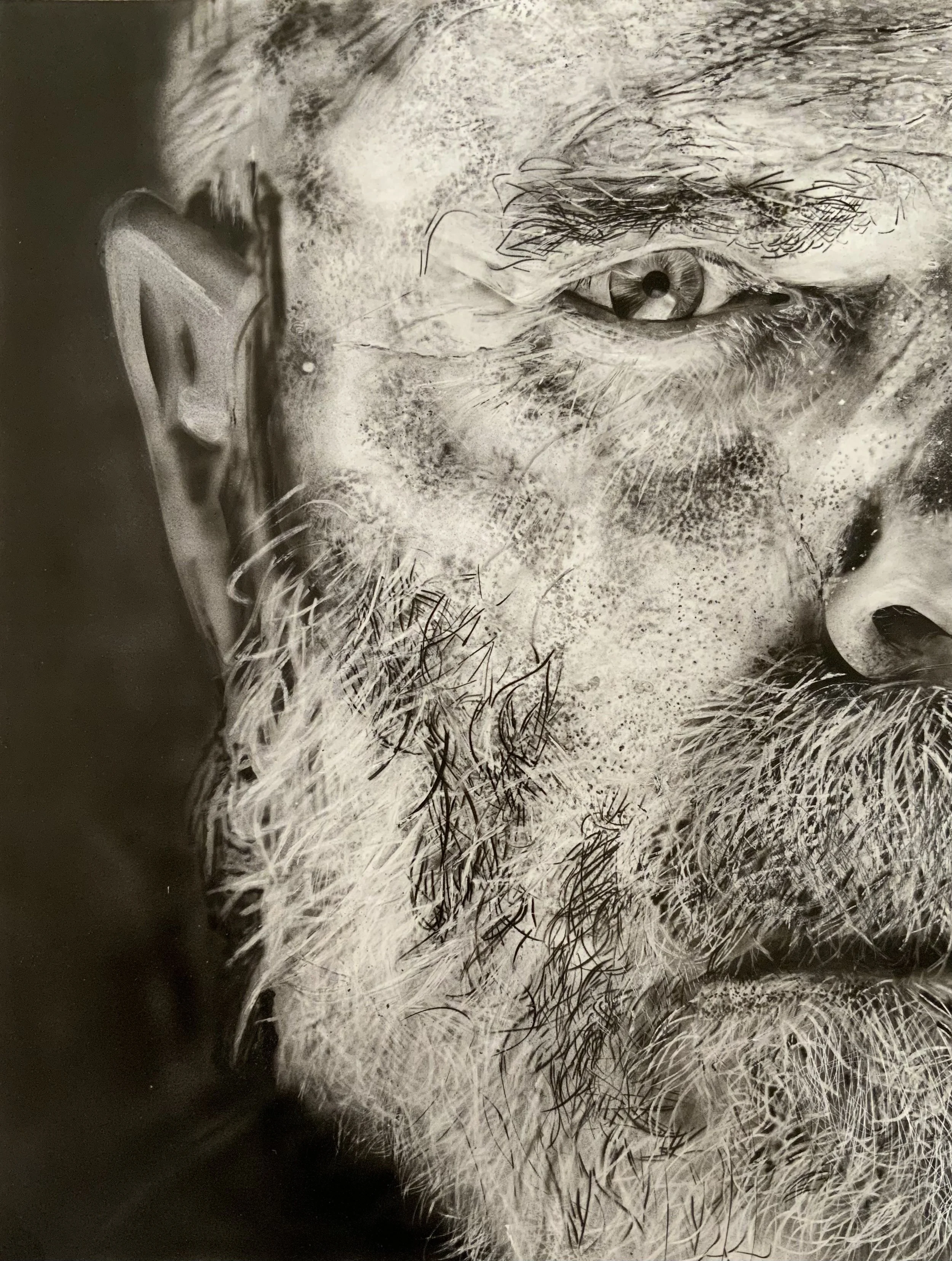 Close-up black-and-white photograph of a person with a beard and mustache, focusing on their eye, eyebrow, and parts of their face and hair.