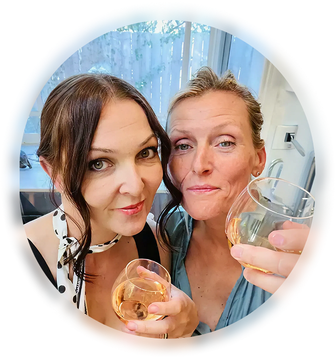 Two women smiling and holding glasses of rosé wine indoors, with a window and blinds in the background.