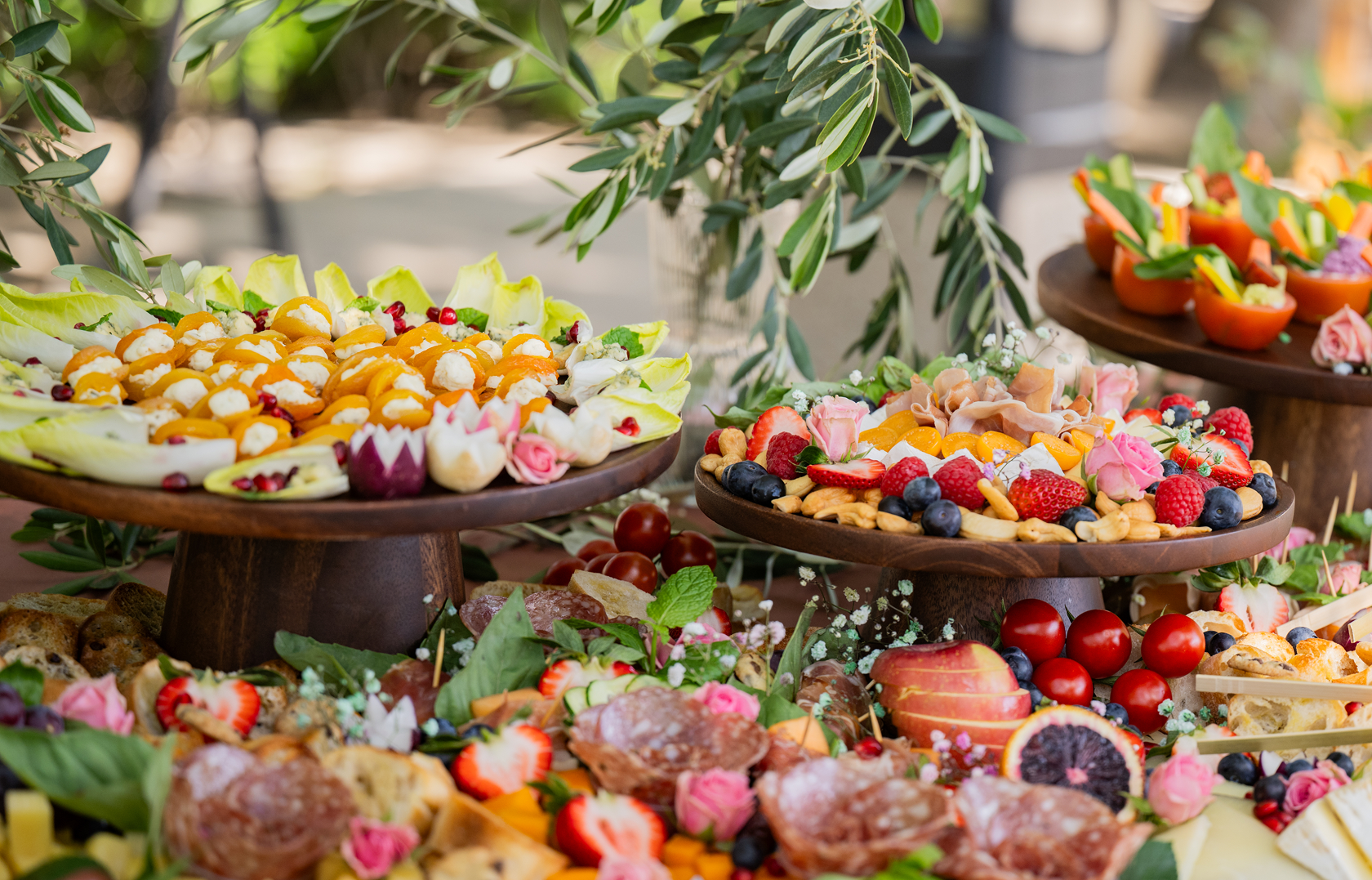 Colorful fruit and cheese platter display with strawberries, blueberries, raspberries, tomatoes, and sliced peaches, decorated with pink roses and green leaves on wooden stands.