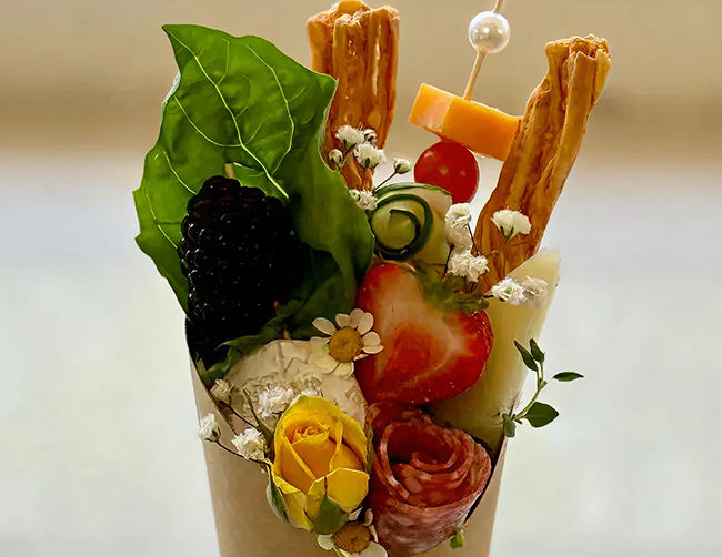 A decorative arrangement of various foods and flowers in a glass, including leafy greens, blackberries, cherry tomatoes, cheese, breadsticks, cucumber slices, and small white and yellow flowers.