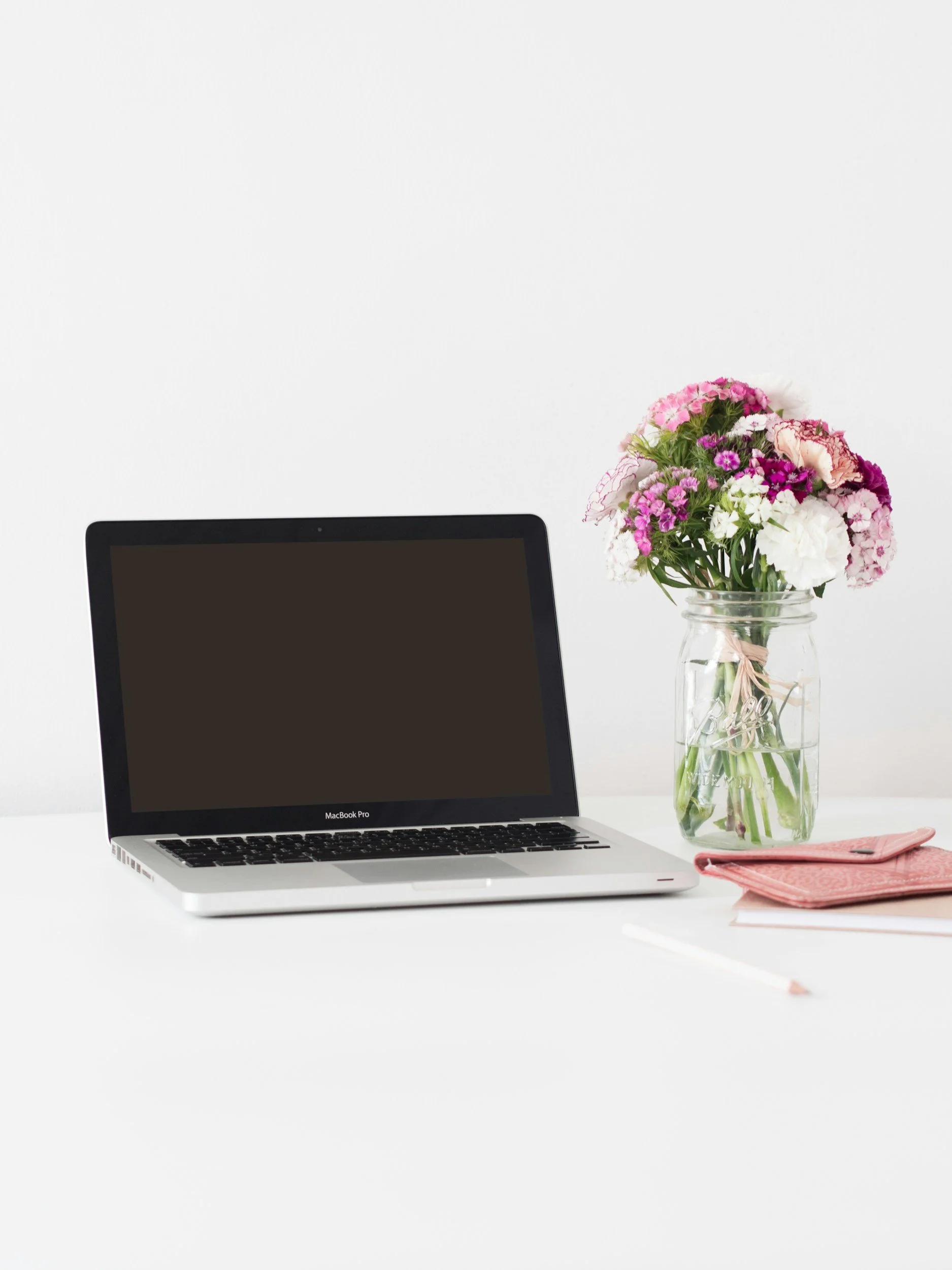 A silver MacBook Pro laptop on a white desk, a pink napkin and pencil in the foreground, and a glass jar filled with pink, white, and purple flowers on the right side, against a plain white background.