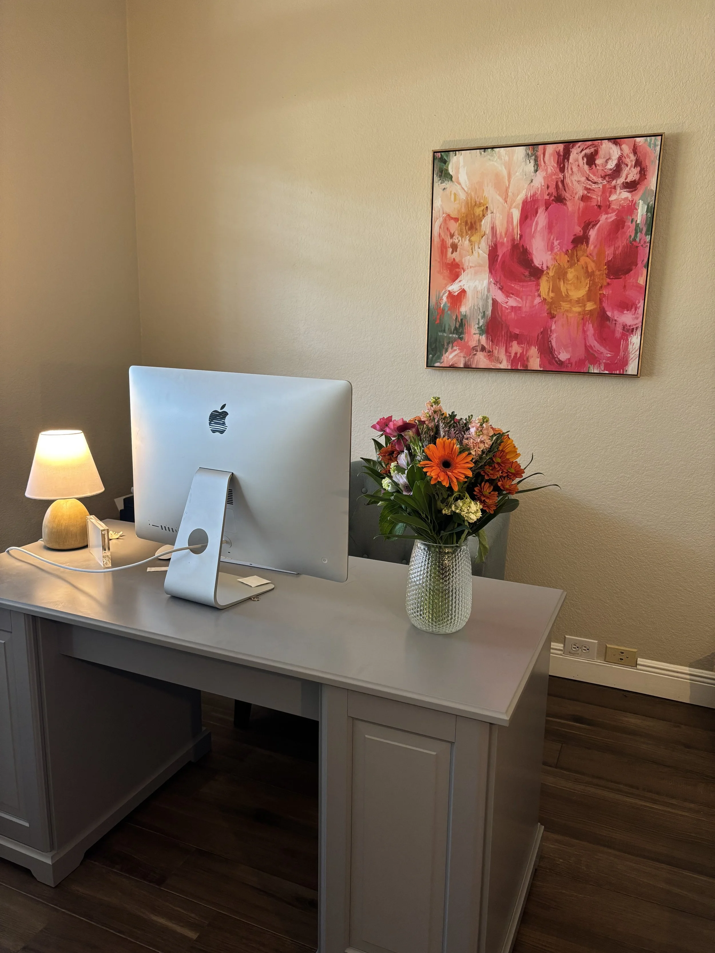 A white desk with a computer monitor, a small lamp, and a vase with a colorful flower arrangement. A painting of pink and red flowers is on the wall behind the desk.