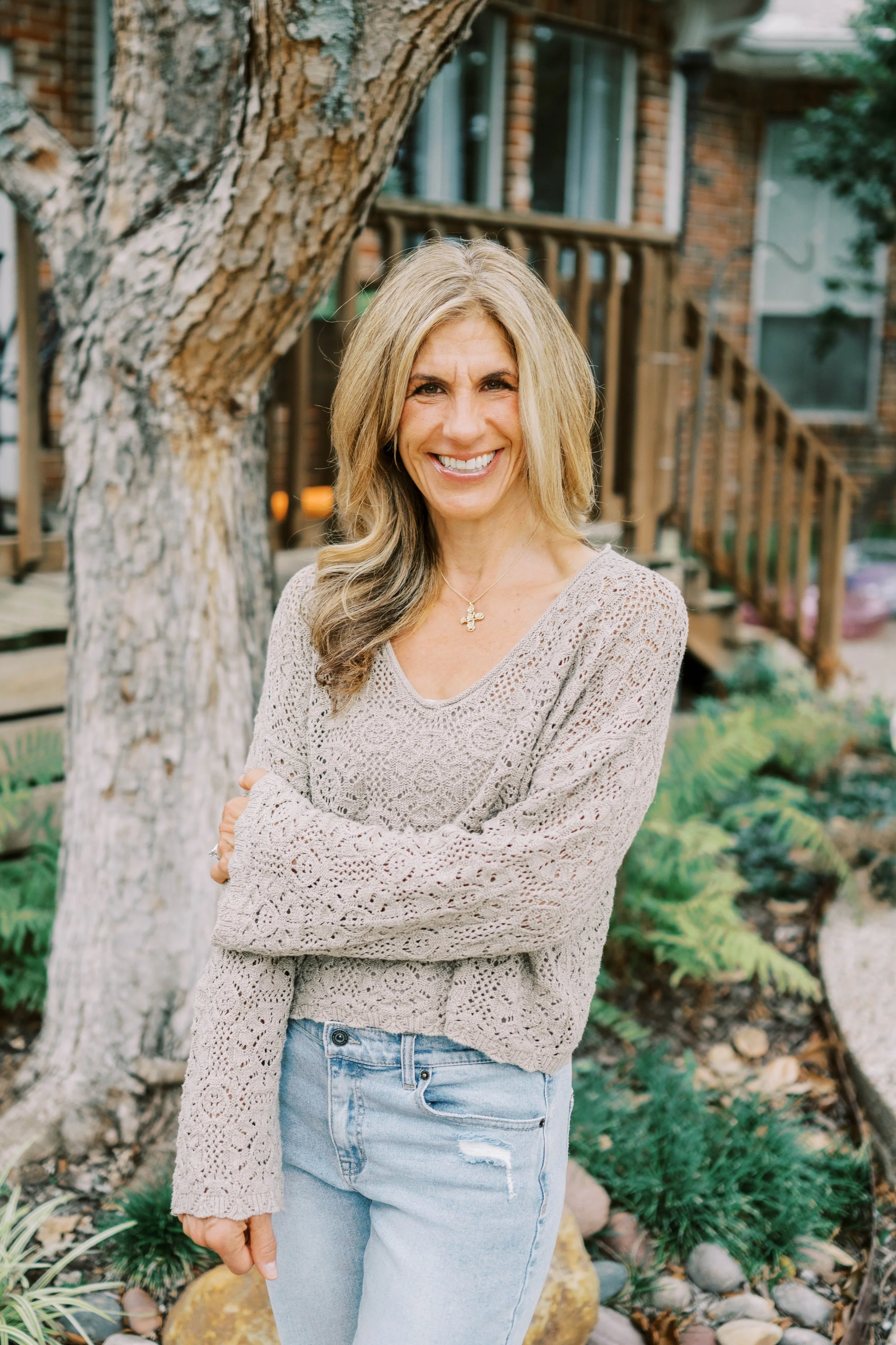 A woman with blonde hair smiling outside in front of a large tree, brick house with stairs, and greenery.