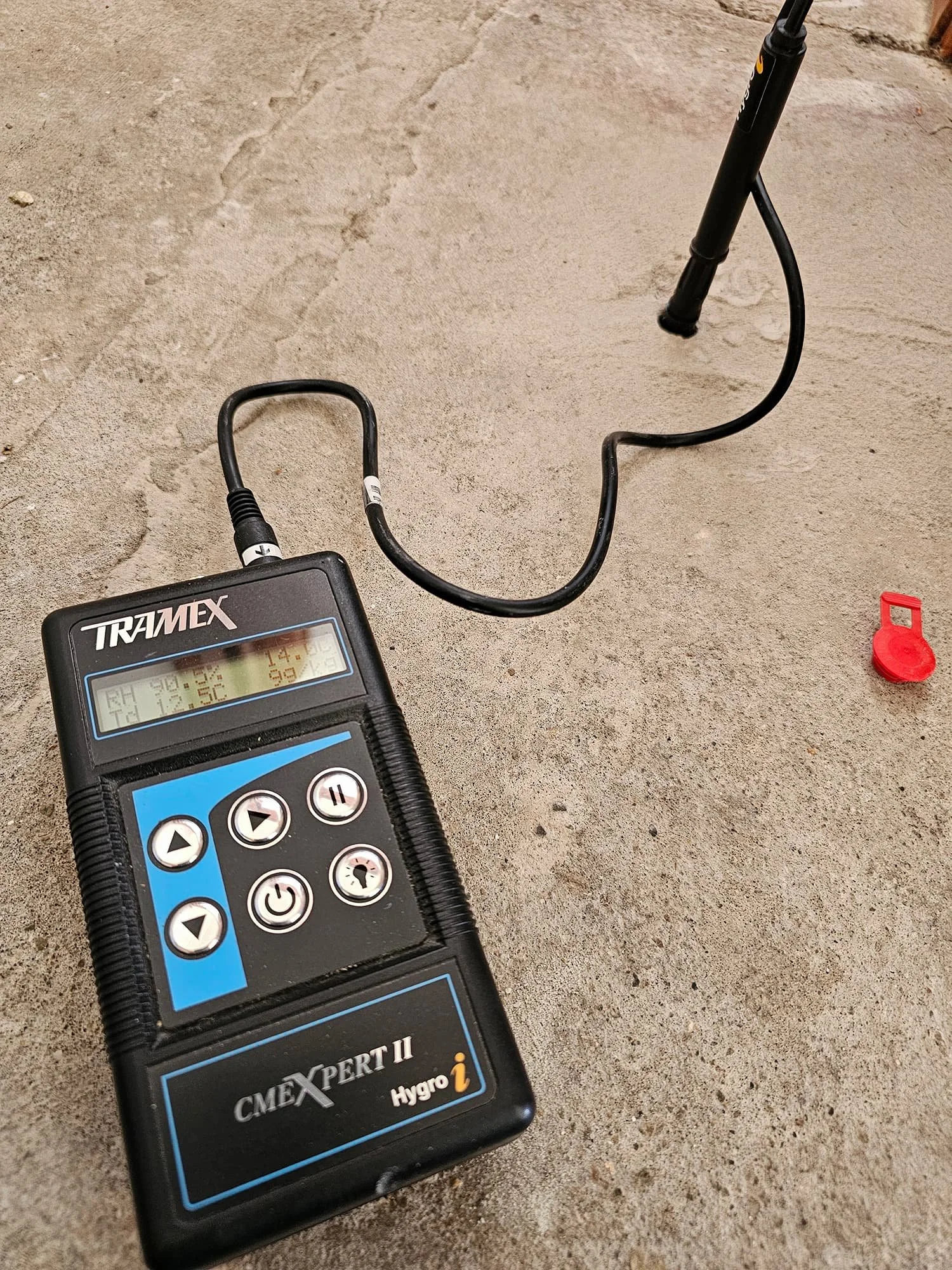 A digital device with a screen displaying some numbers connected to a black cable. The device reads 14.2, 99, 12.52, and other numbers. The device is labeled TRAMEX and CMEXPERT II. It is on a concrete surface, with a red object and some other small items nearby.