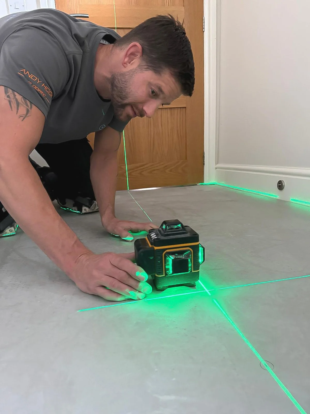 Man kneeling on the floor using a laser level to project green laser lines onto the floor and walls for leveling or alignment.