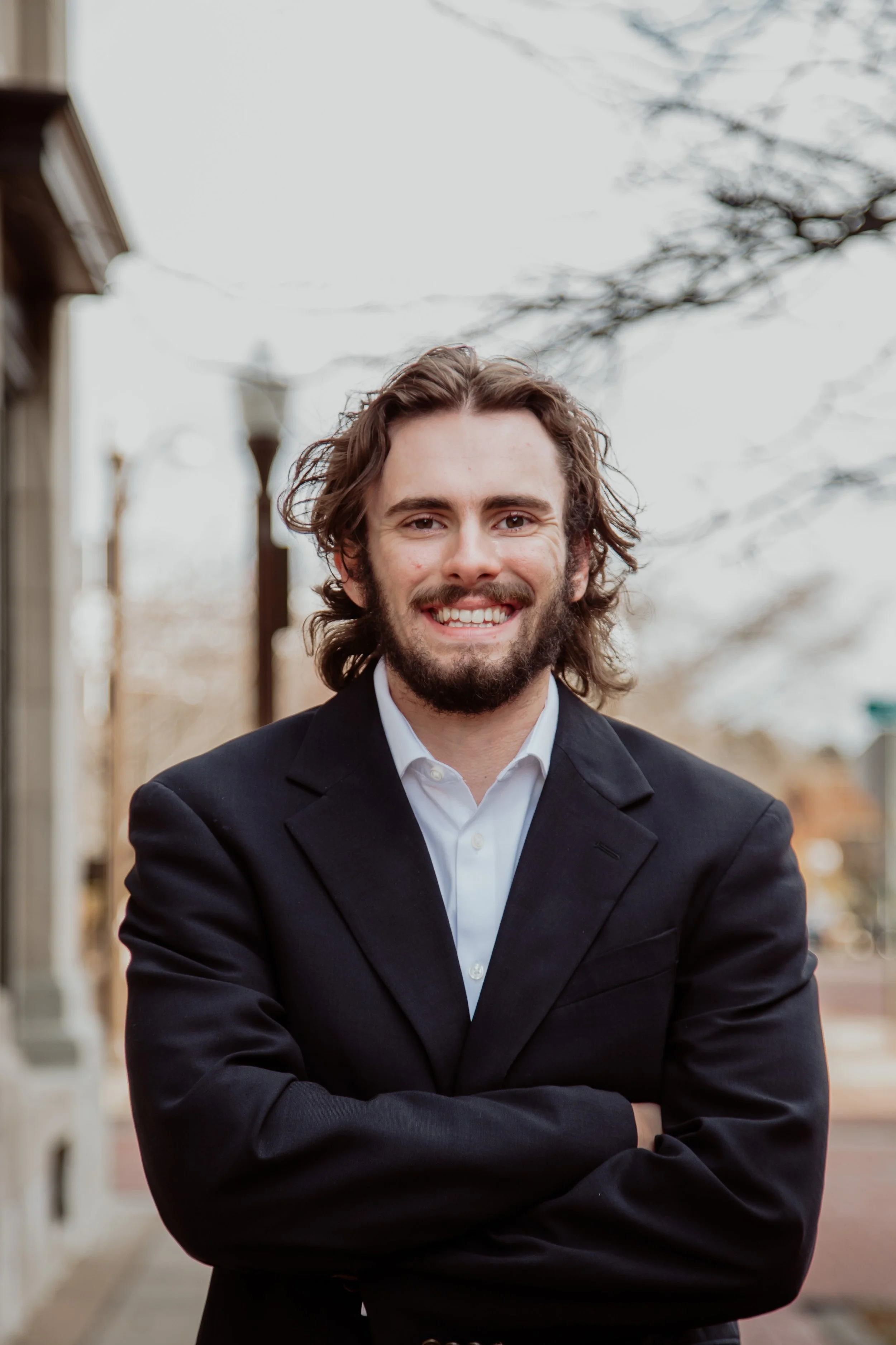 A smiling man with brown hair, a beard, wearing a light blue dress shirt and a dark blazer, standing against a plain background.