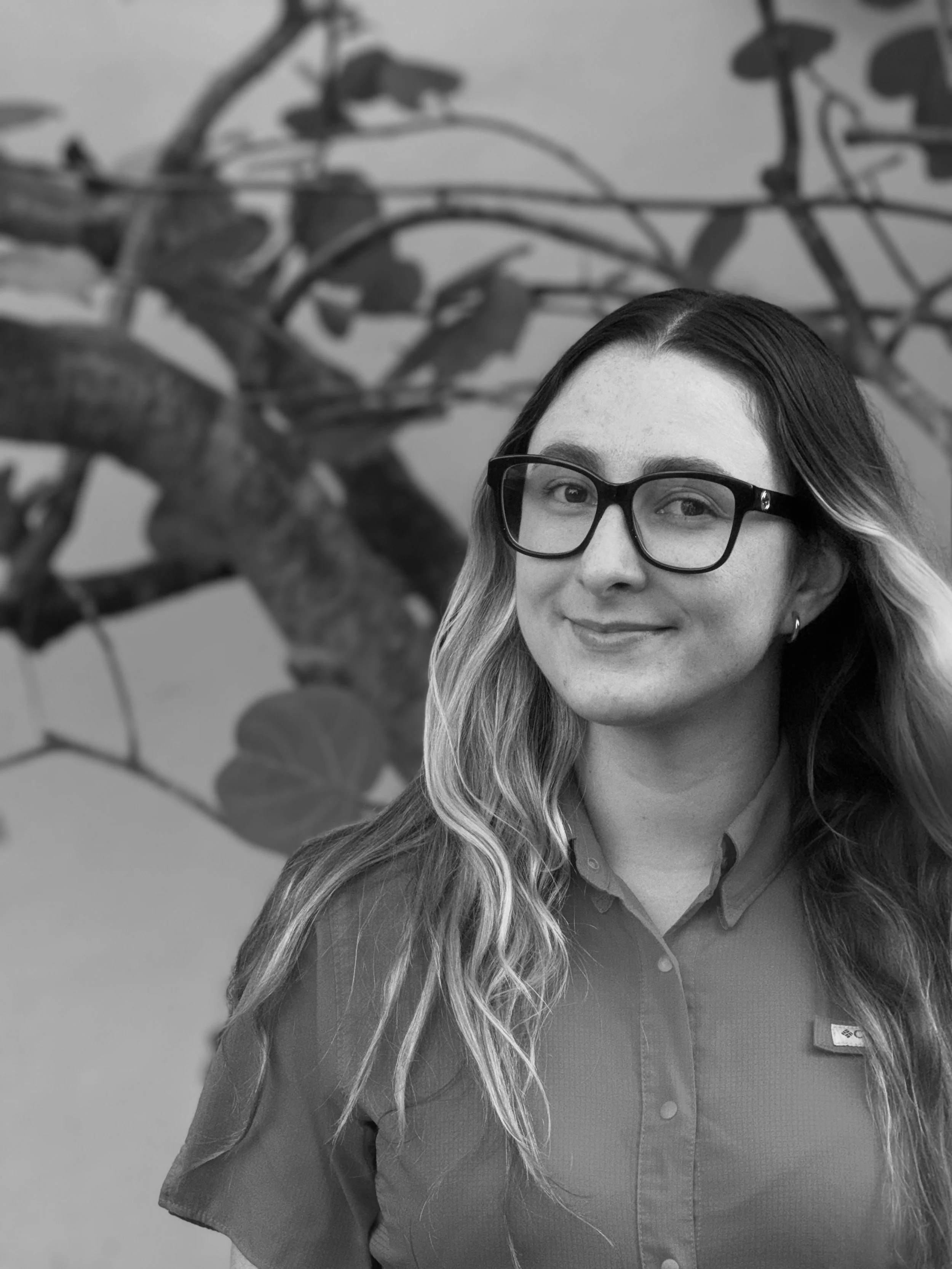 Illustration of a young woman with long blonde hair, round glasses, and a small earring, smiling and wearing a cream-colored shirt.