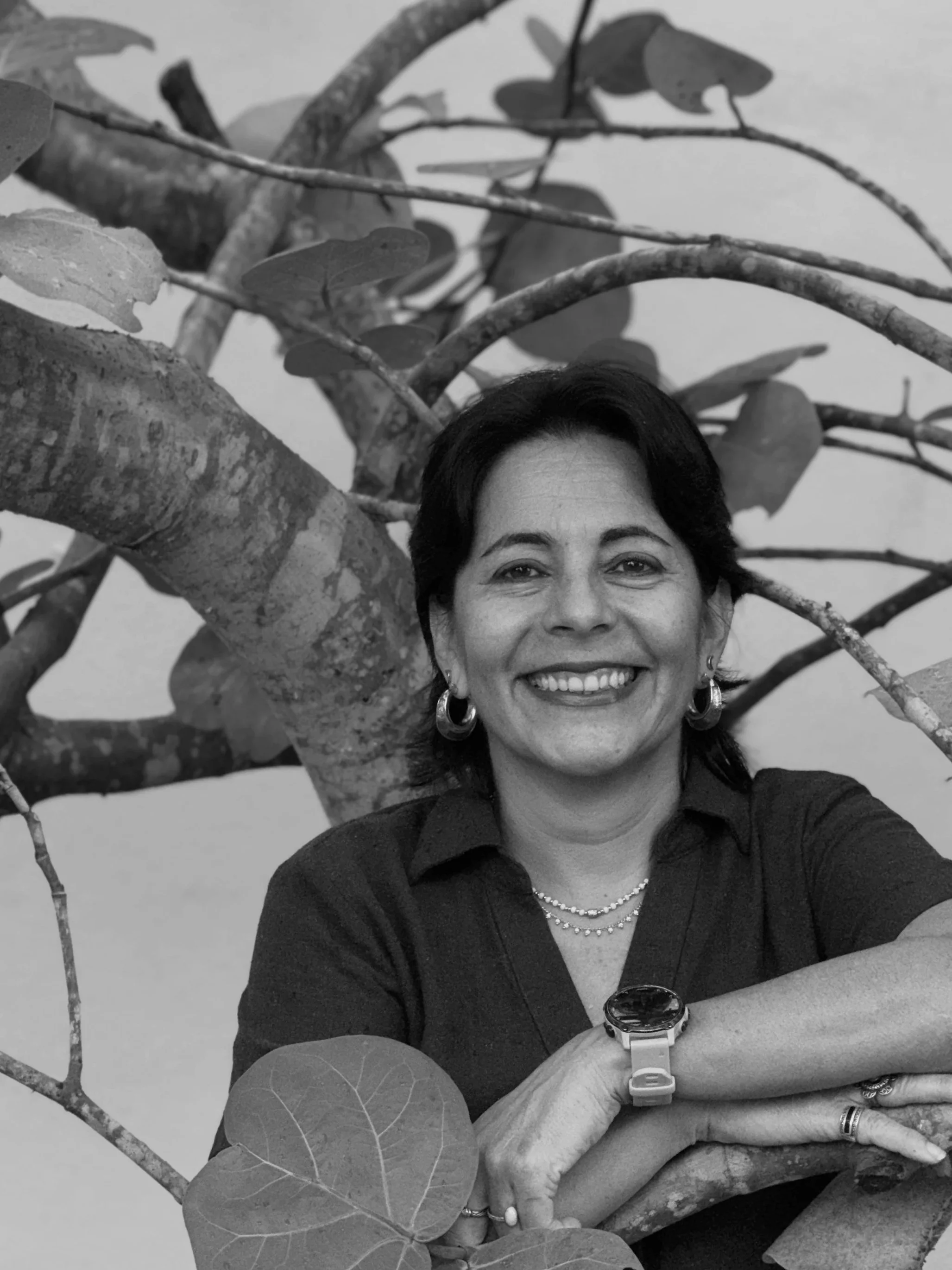 An animated woman with brown hair tied back in a ponytail, wearing black sunglasses, a green shirt, and a broad smile, standing outdoors with the ocean and a partly cloudy sky in the background.