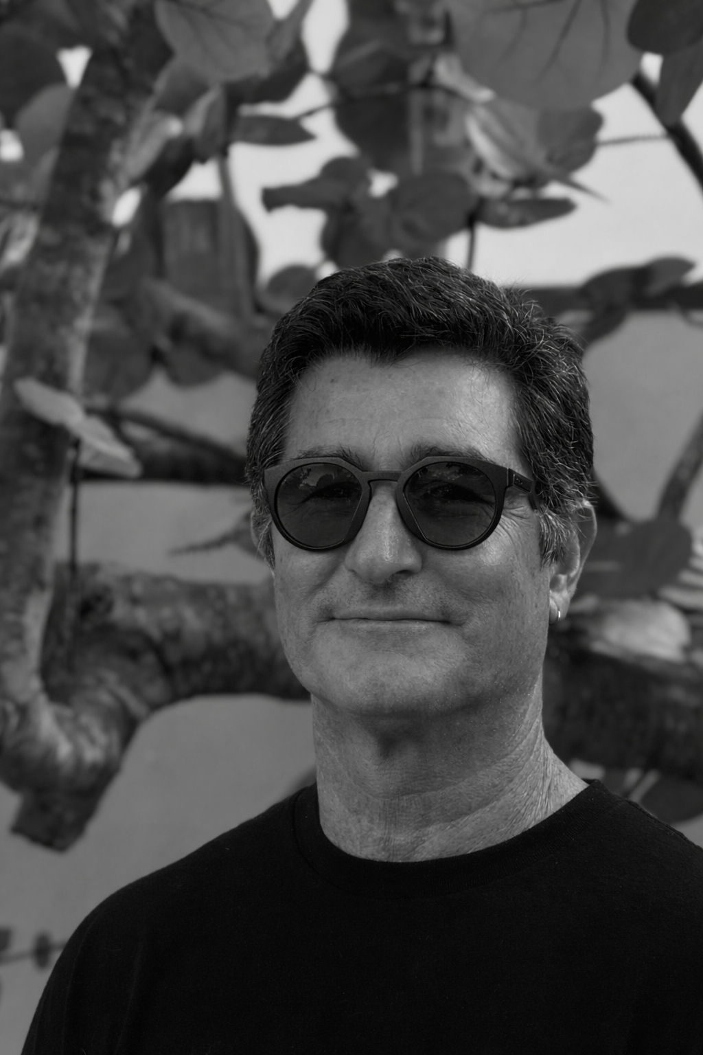 An animated woman with brown hair tied back in a ponytail, wearing black sunglasses, a green shirt, and a broad smile, standing outdoors with the ocean and a partly cloudy sky in the background.