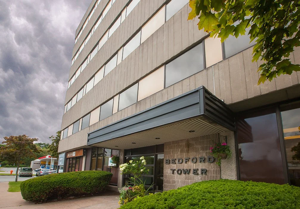 Exterior view of a modern multi-story office building named Bedford Tower, with bushes, trees, and a parking lot in front, under a cloudy sky.