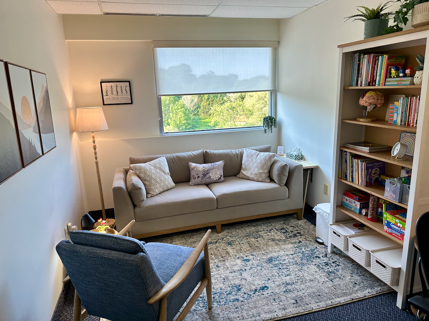 Cozy living room with a beige sofa with patterned pillows, a blue armchair, a floor lamp, a bookshelf filled with books and decor, a window with greenery outside, and a decorative rug.