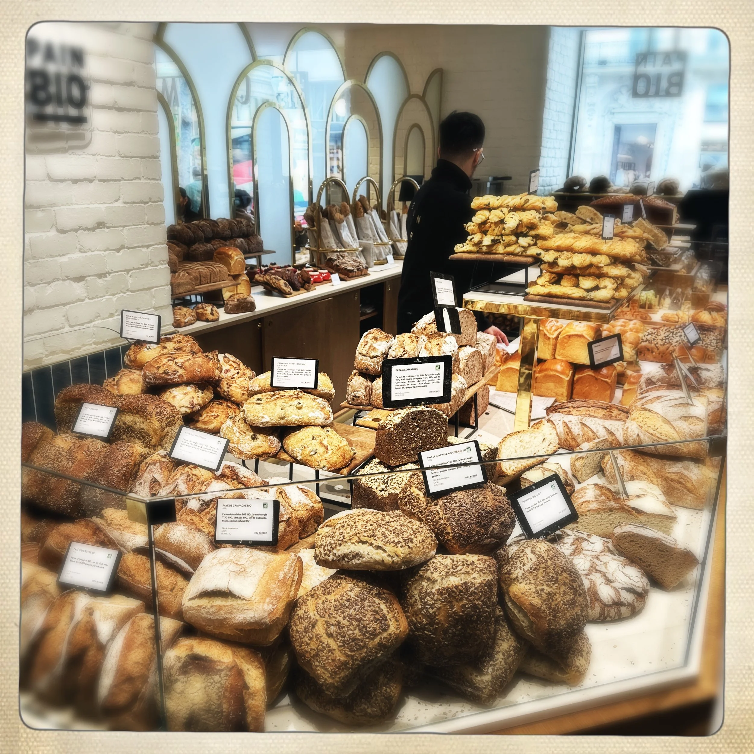 Display of various breads in a Parisian bakery with a worker behind the glass and a background of windows.