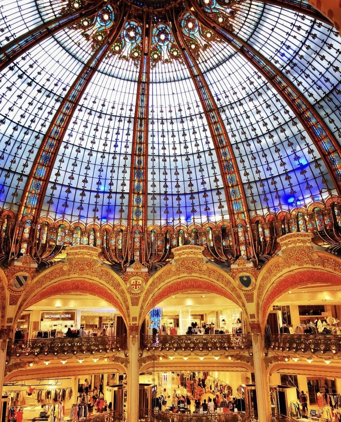 Interior view the large stained glass dome ceiling at Galeries Lafayette in Paris, multiple floors with shops, and ornate architectural details.