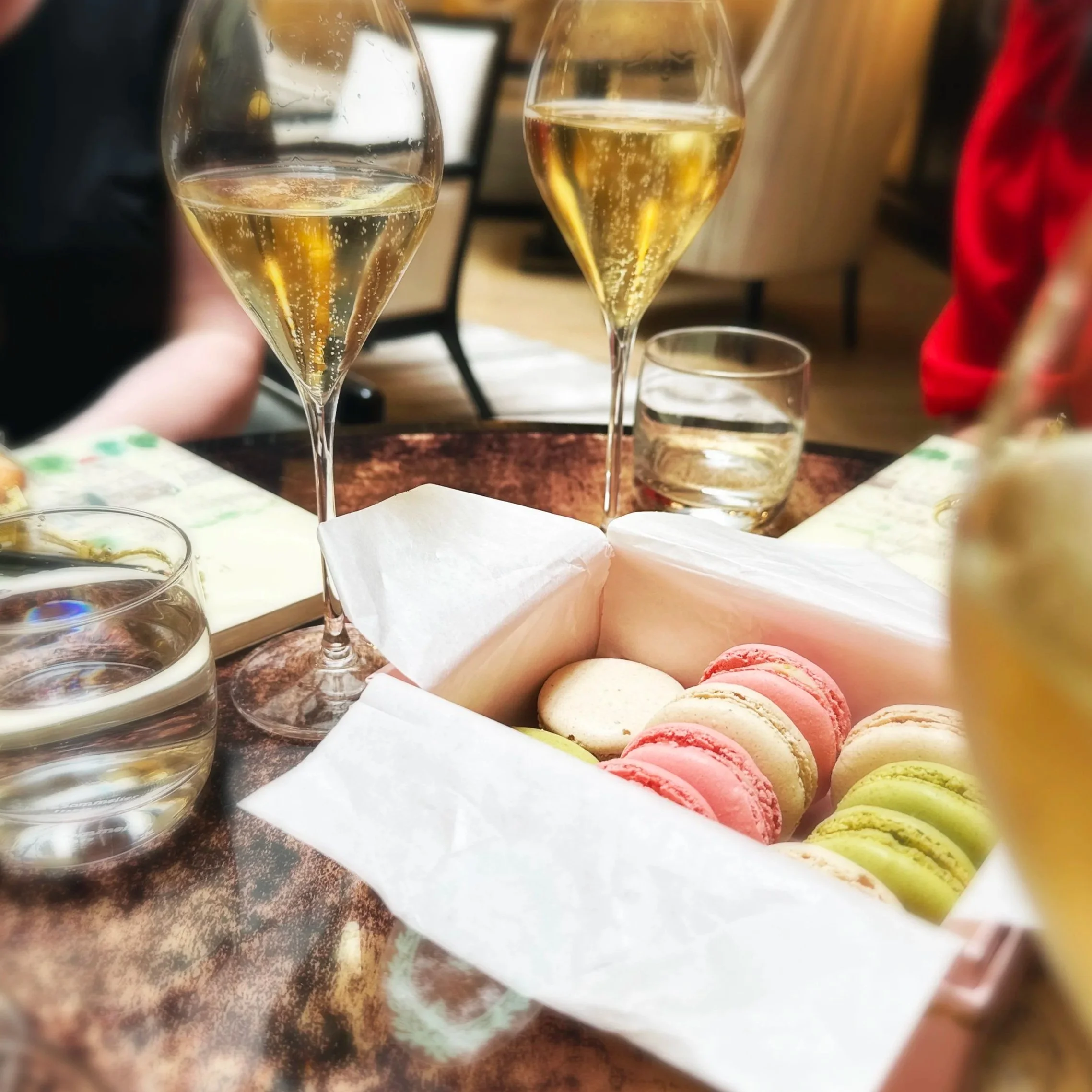 Two glasses of white wine, a glass of clear liquid, a box of colorful macarons, and glasses of water on a table in a cozy setting.