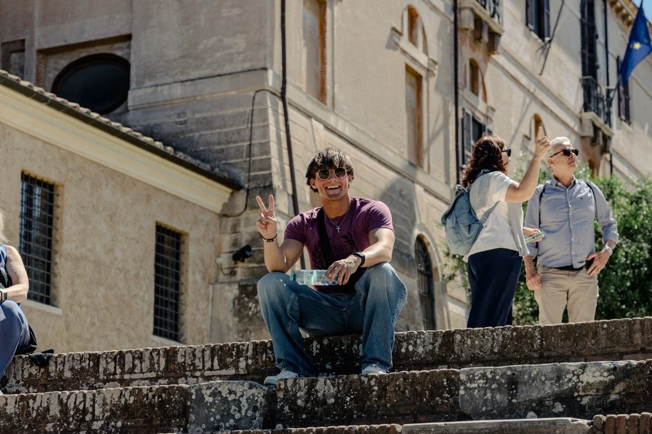A young man wearing sunglasses sitting on stone steps, smiling, making a peace sign with his right hand, and holding a water bottle in his left hand. Behind him, two women are talking, one of whom is pointing upwards. The scene is set outdoors with historic buildings in the background.