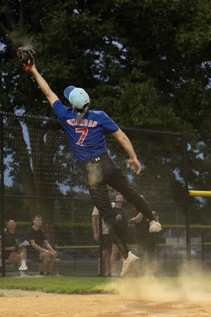 A baseball player wearing a blue jersey with the number 7 and the name Swanson is jumping to catch a ball with a glove during a game.