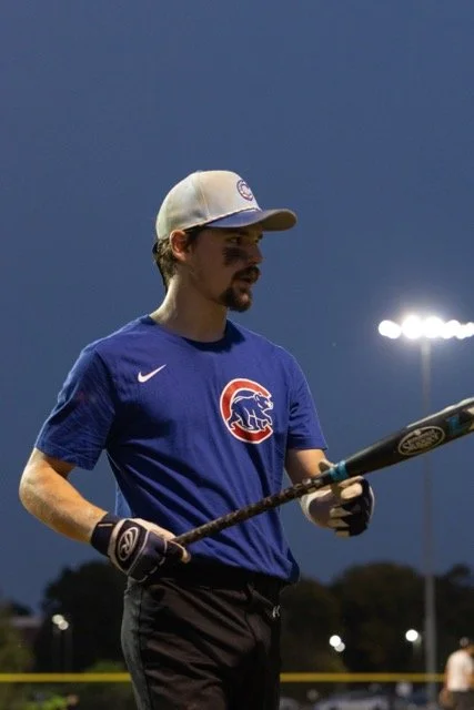 A man in a blue Chicago Cubs baseball jersey and a baseball cap holding a baseball bat on a baseball field at night.