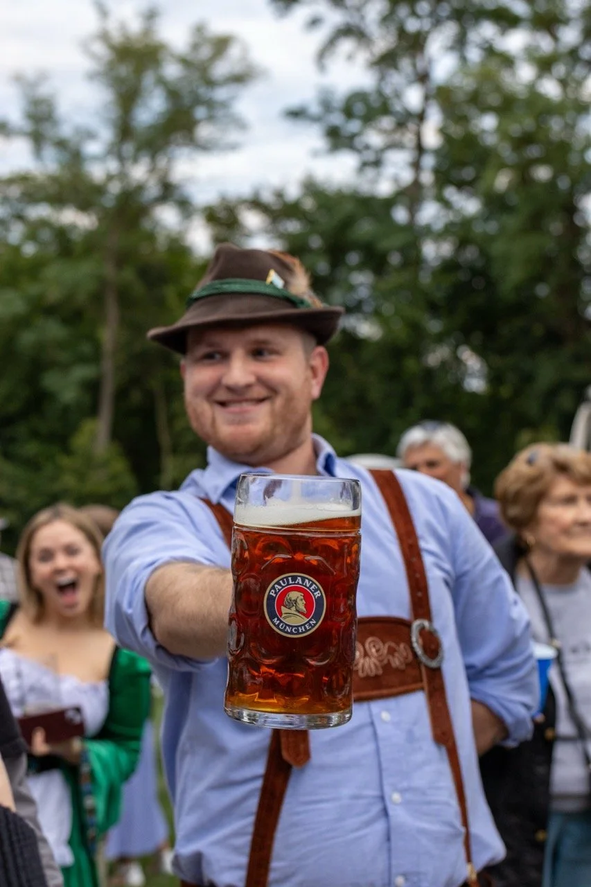 A man in traditional Bavarian attire holding a large glass of beer at an outdoor gathering, with other people in festival clothing in the background, surrounded by trees.
