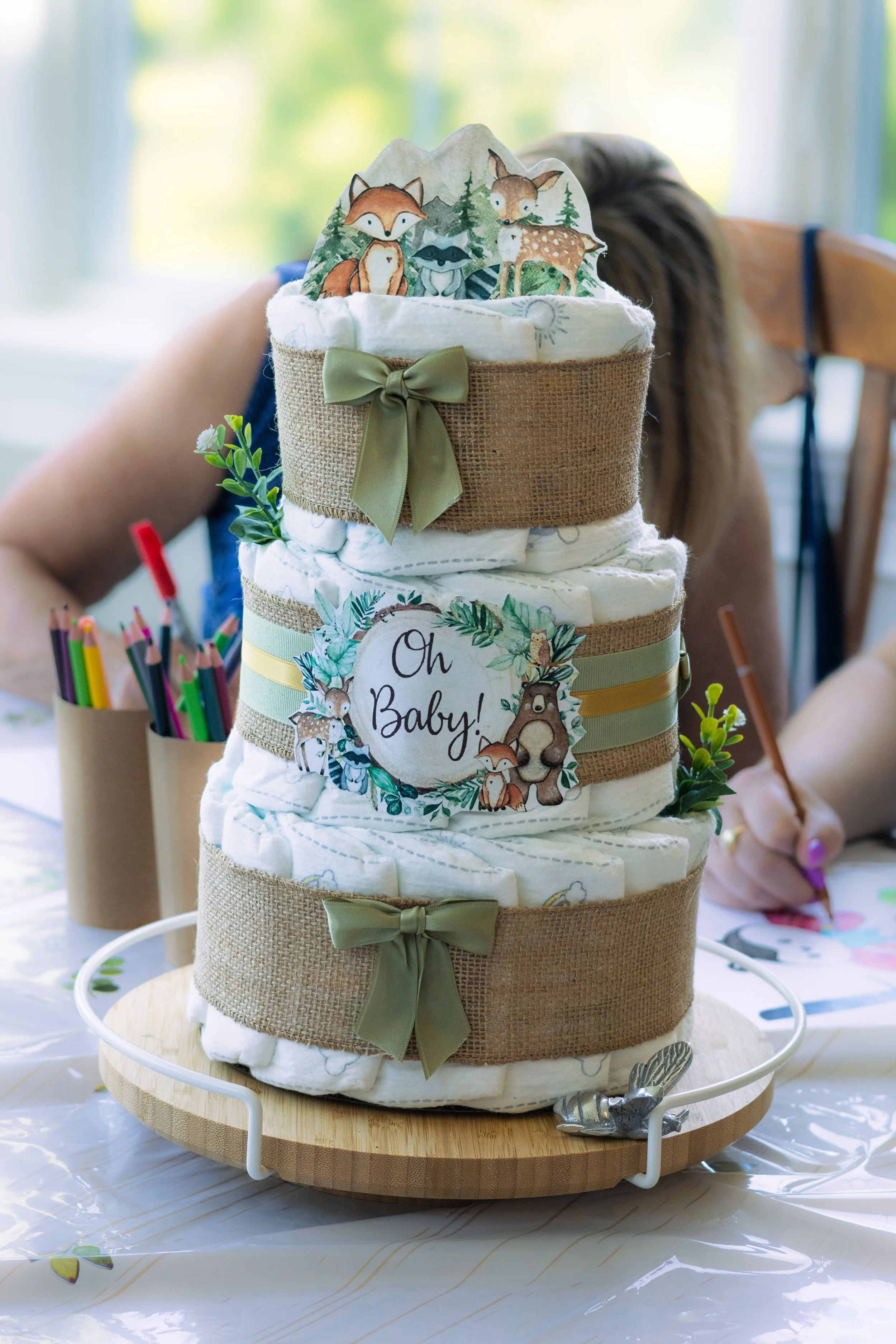 A layered diaper cake decorated with woodland animal theme and green bows, featuring a tag that says "Oh Baby!". In the background, a woman sits at a table with colored pencils and drawing supplies.