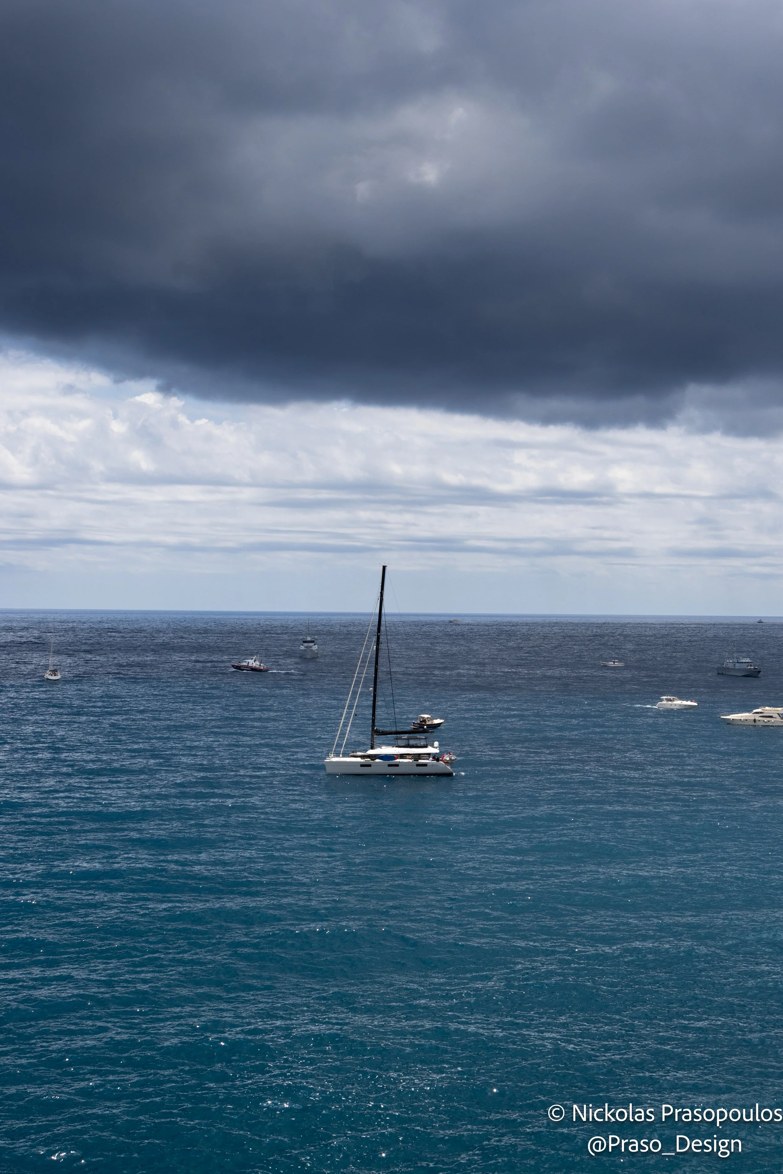 A view of several boats anchored in a calm ocean under a cloudy sky with dark clouds overhead.