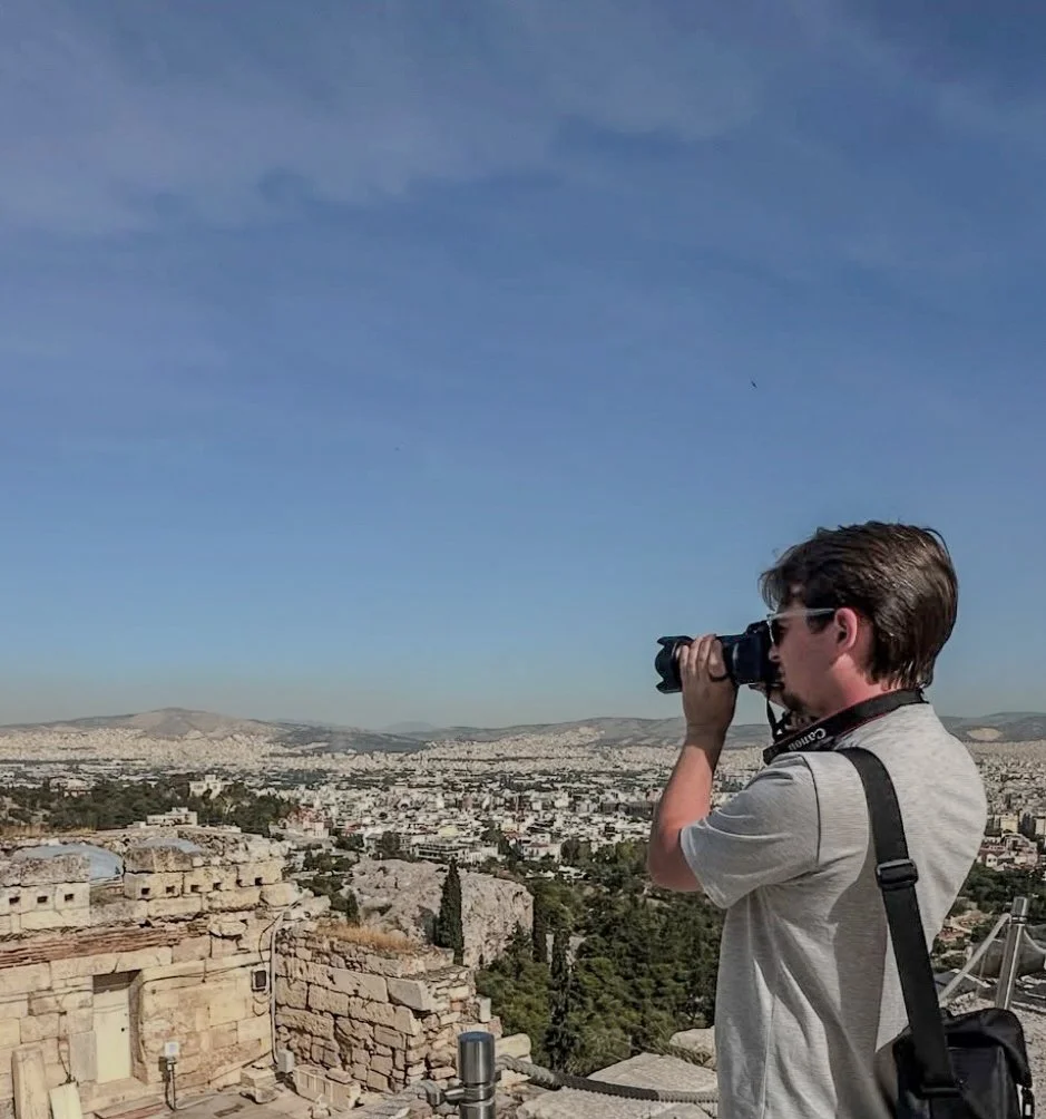 Man taking a photograph with a camera on a historic site overlooking a cityscape with hills in the distance.