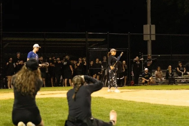 A softball game at night with players and spectators. A woman is preparing to bat, while others are watching from behind the fence and sitting on benches.