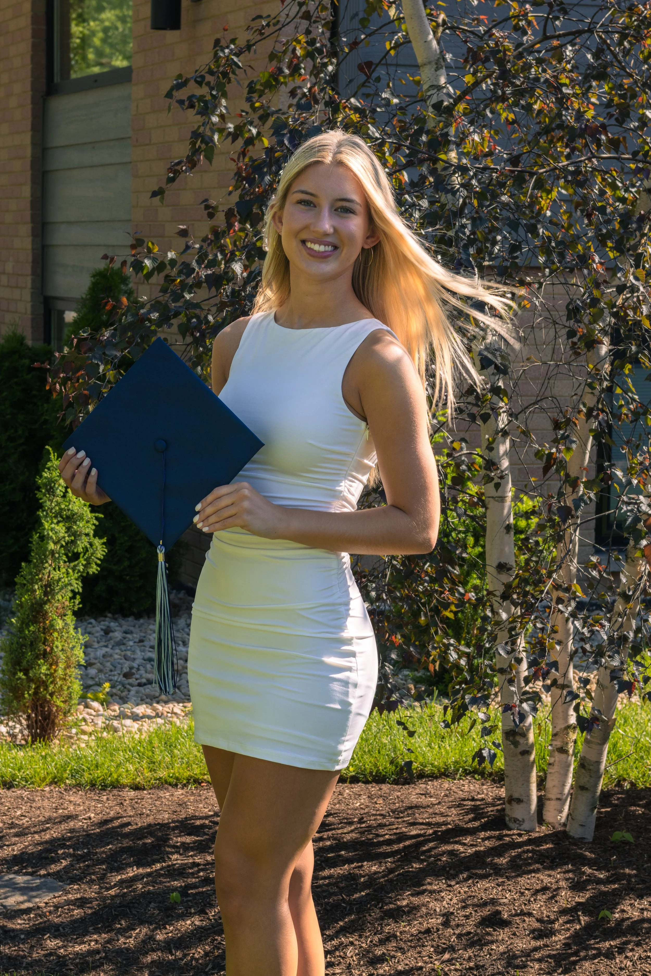 A young woman with blonde hair smiling and holding a graduation cap outdoors in front of a tree and modern building, on a sunny day.