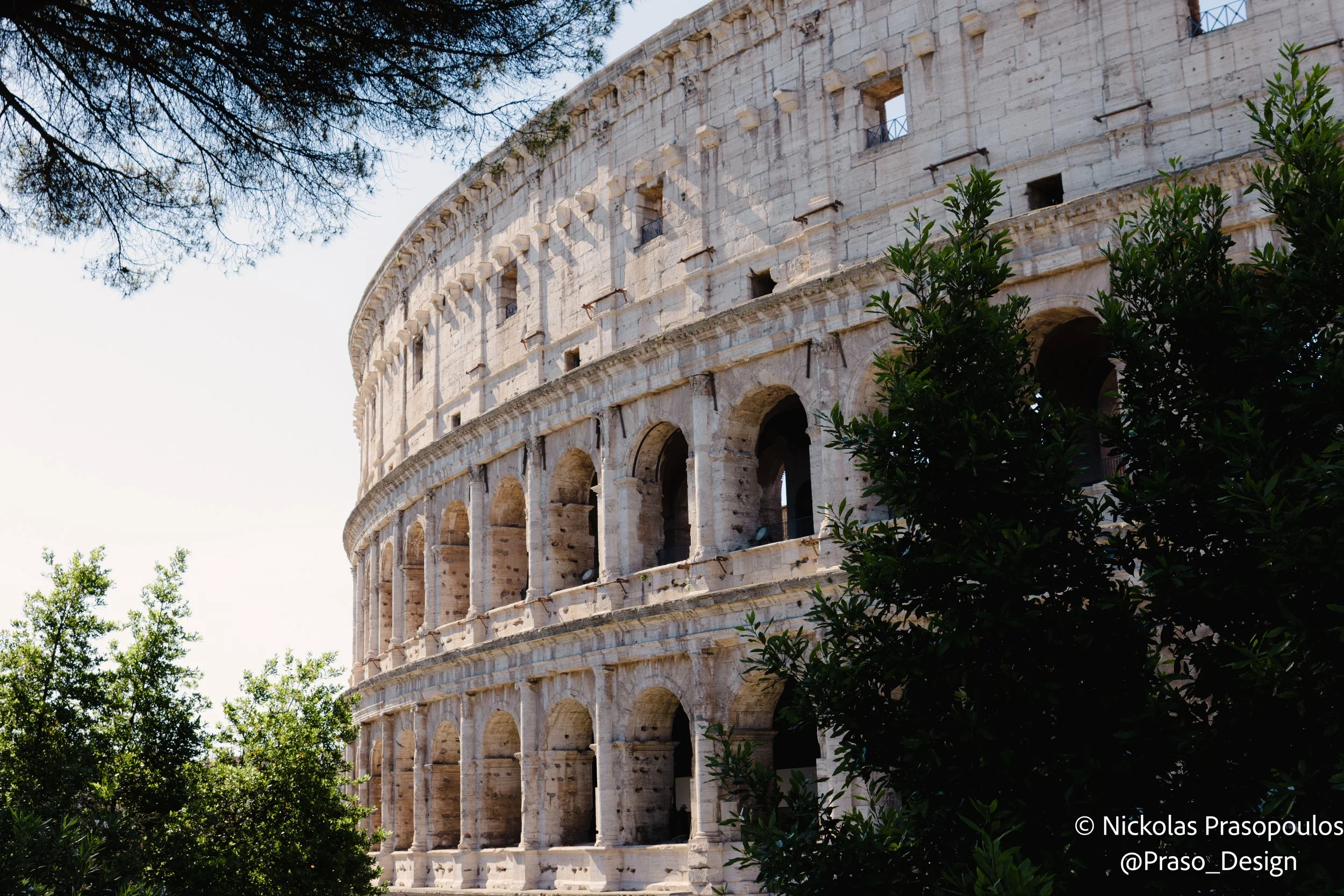 The image shows the exterior of the Roman Colosseum, an ancient stone amphitheater with multiple arches on each level, partially obscured by green trees, under a clear sky.