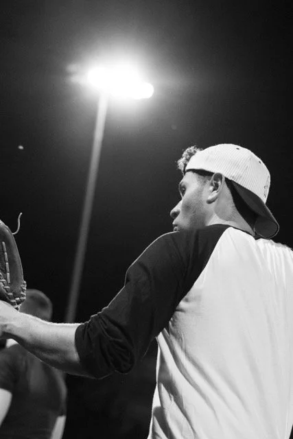 A young man wearing a baseball cap backwards, seen from the side, at night with a bright light overhead, possibly in a sports or outdoor setting.