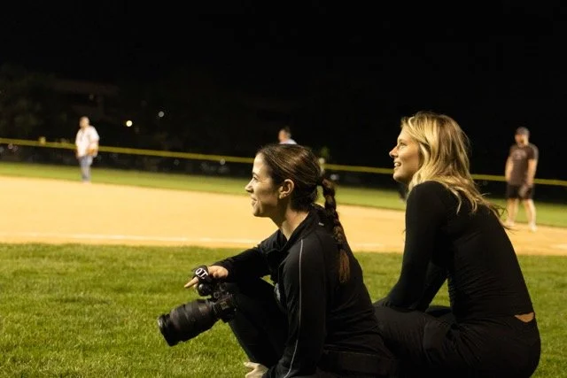 Two women sitting on grass at a baseball field at night, watching a game, with other spectators in the background.