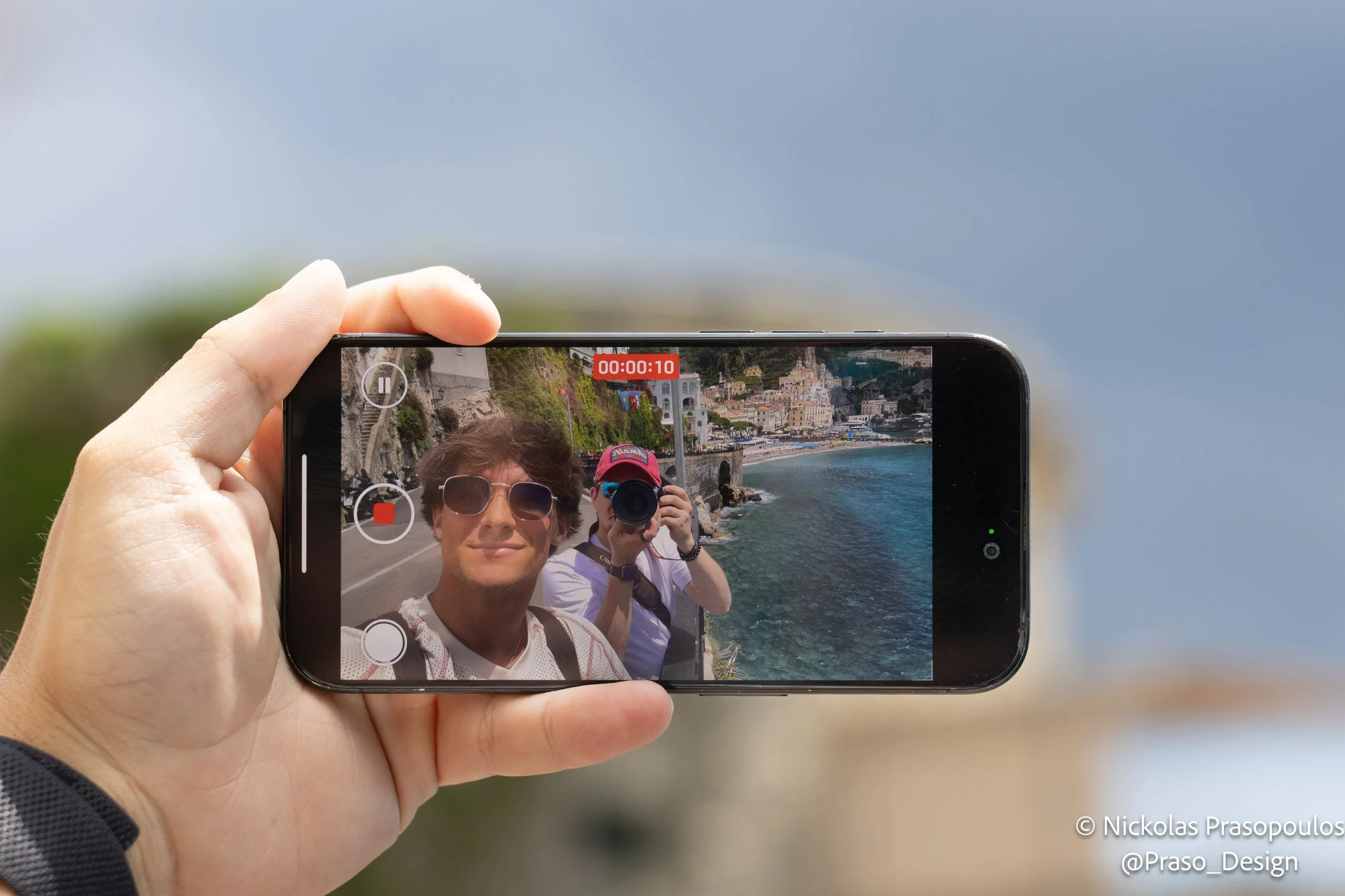Person holding smartphone taking a selfie with two others by a seaside town in the background.