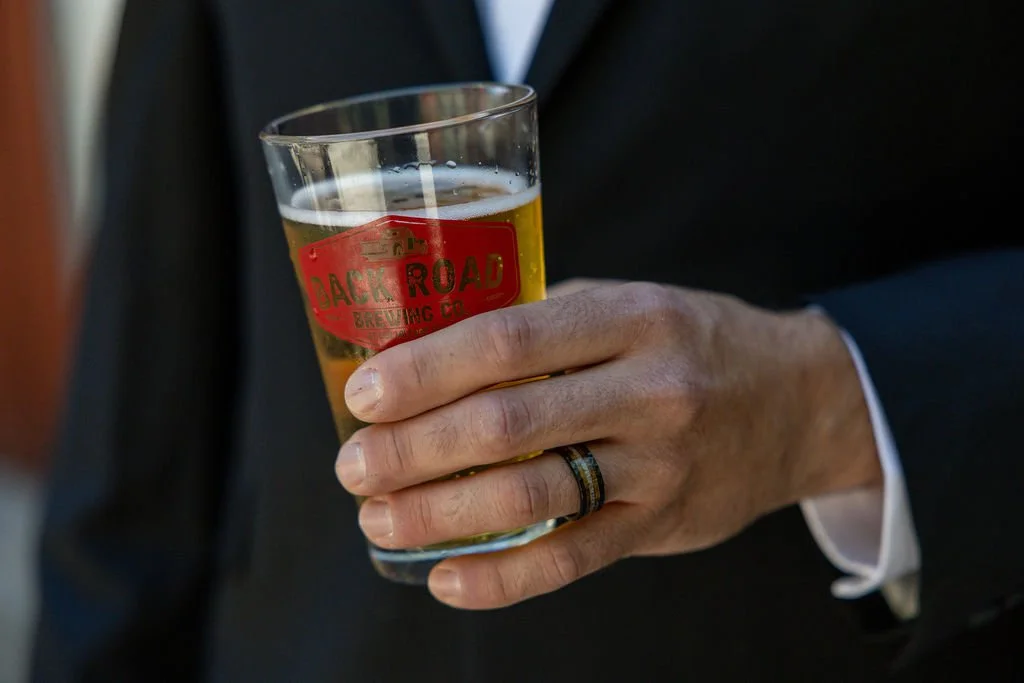 Close-up of a person in a suit holding a glass of beer with a "Back Road Brewing Co" logo.