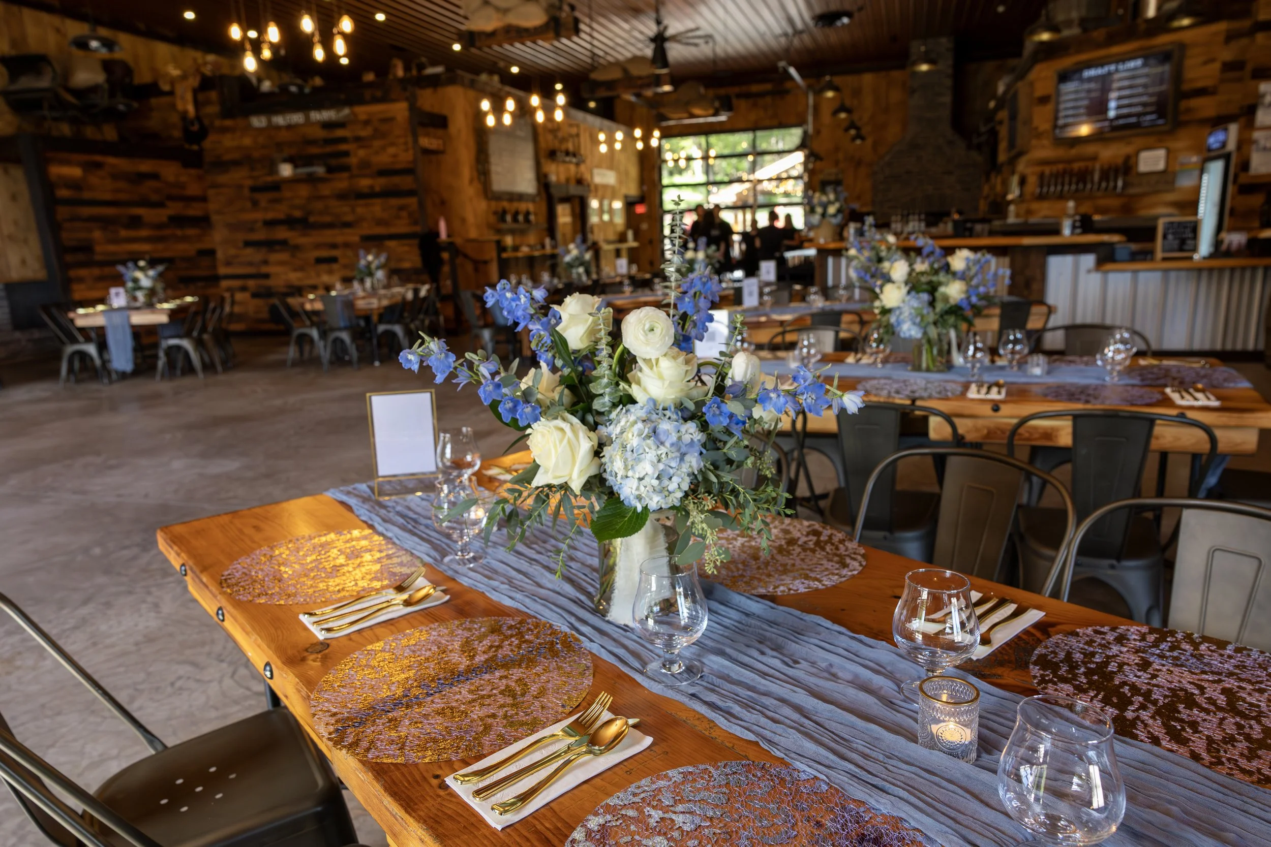 Interior of a rustic restaurant decorated for an event with wooden tables, blue and white floral centerpieces, gold flatware, and decorative placemats.