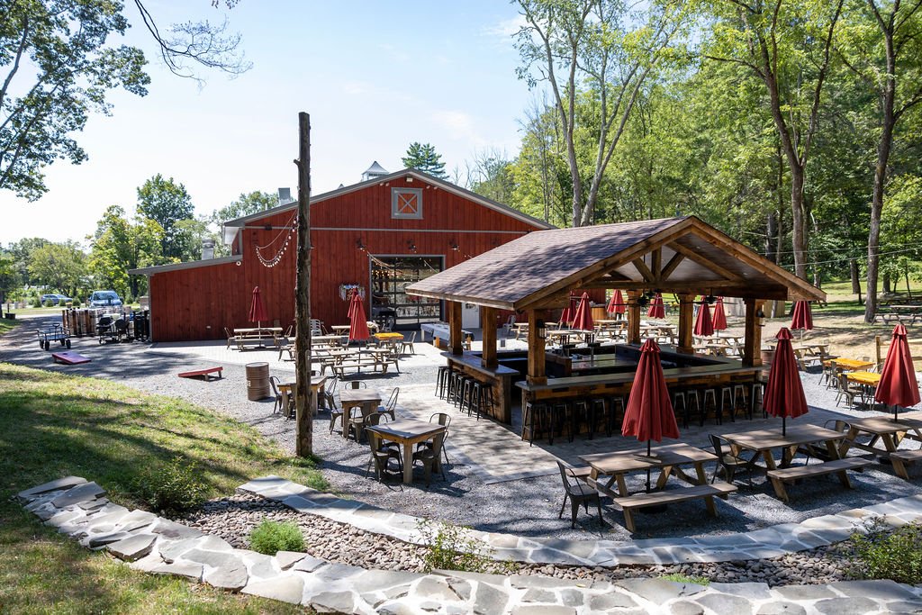 An outdoor patio area with wooden picnic tables and red umbrellas, adjacent to a red barn-style building in a wooded area on a sunny day.