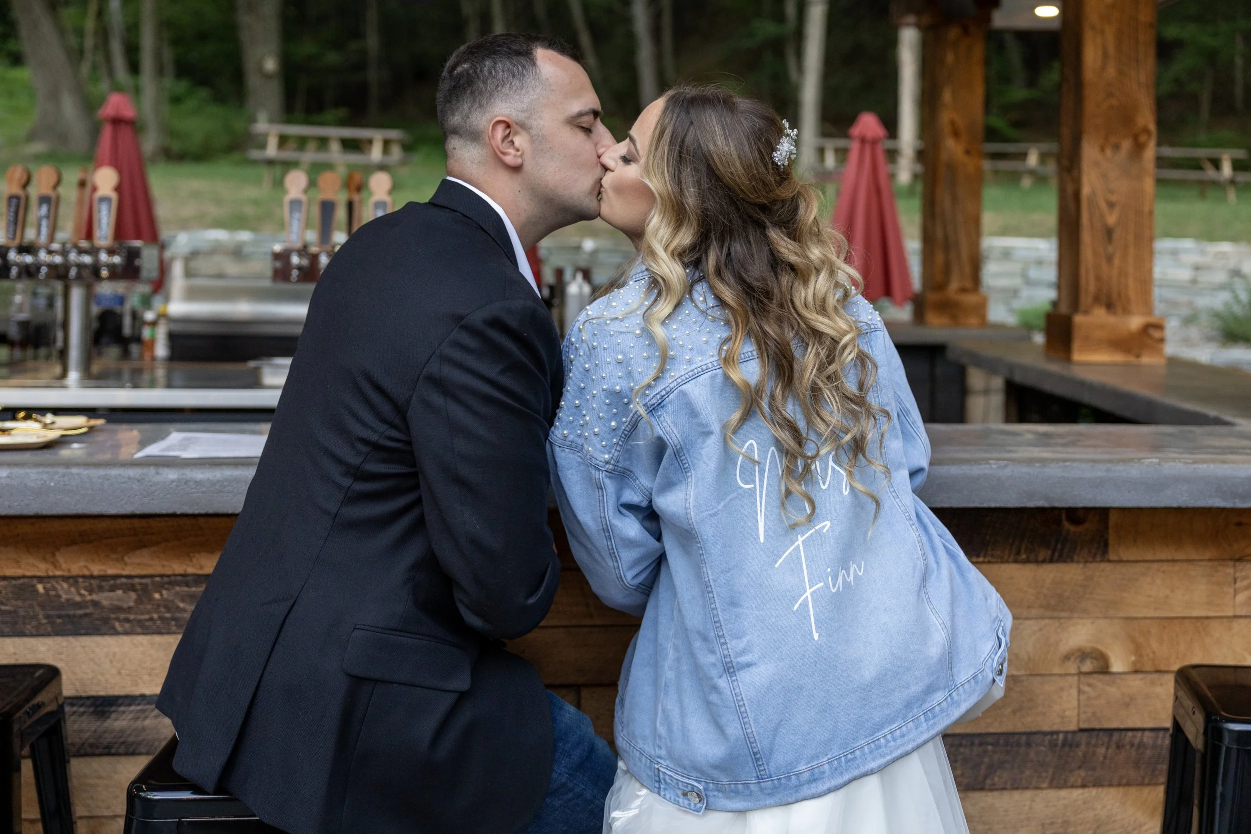 A couple sharing a kiss at an outdoor bar, with the woman wearing a denim jacket and the man in a black suit, in a wooded setting.