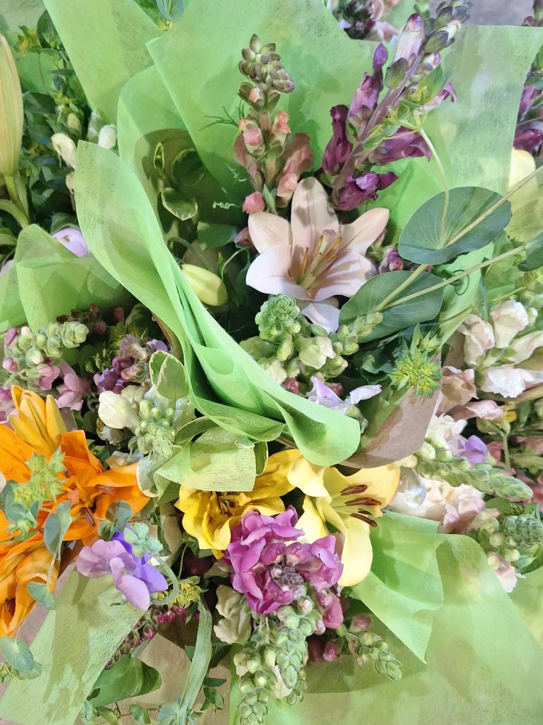 A colorful bouquet of various flowers including lilies, snapdragons, and other blooms, wrapped in green paper and brown craft paper.