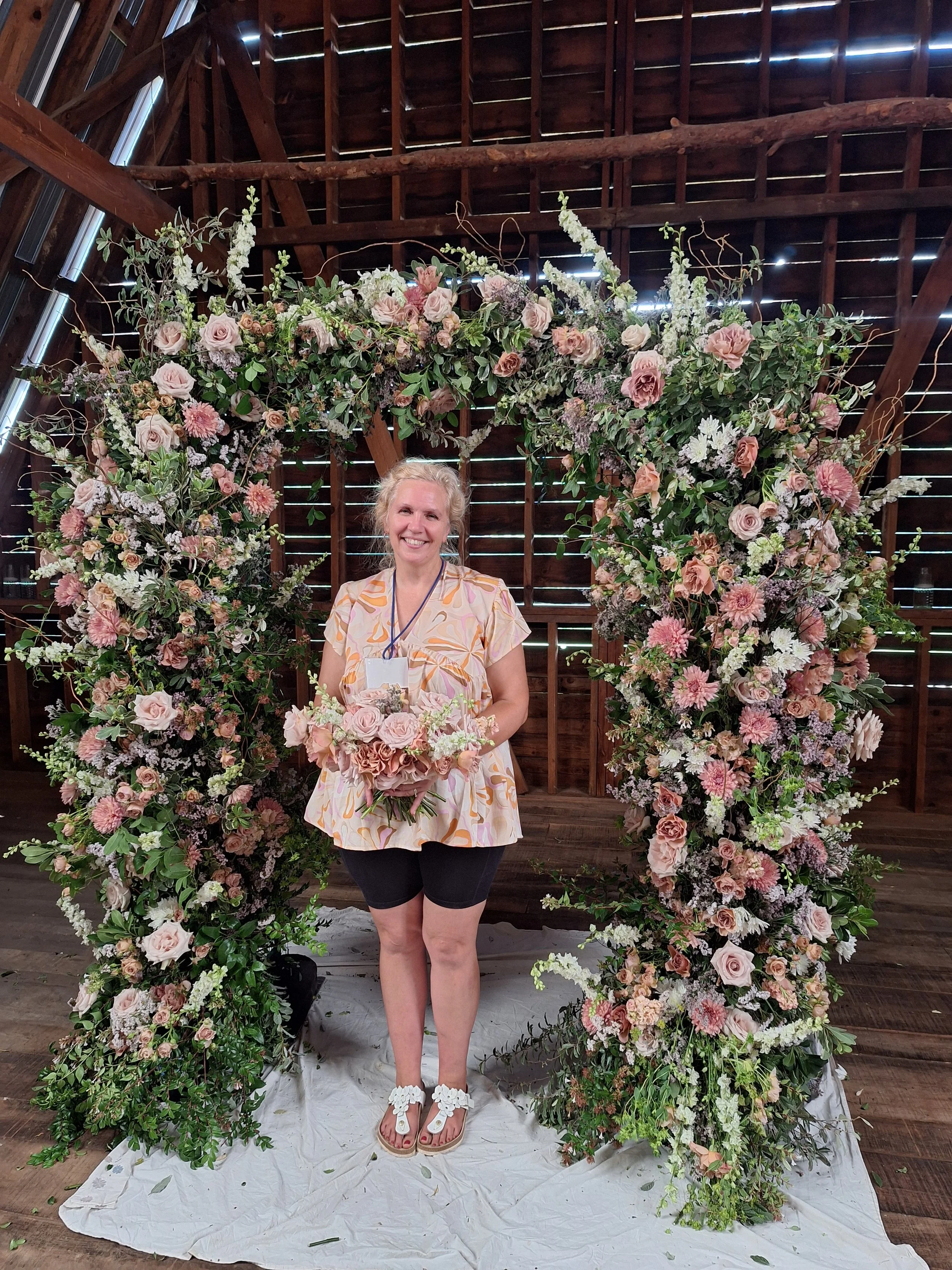 A woman standing in front of a large floral arch, holding a bouquet of pink and white flowers, inside a wooden barn with sunlight filtering through gaps in the wall.
