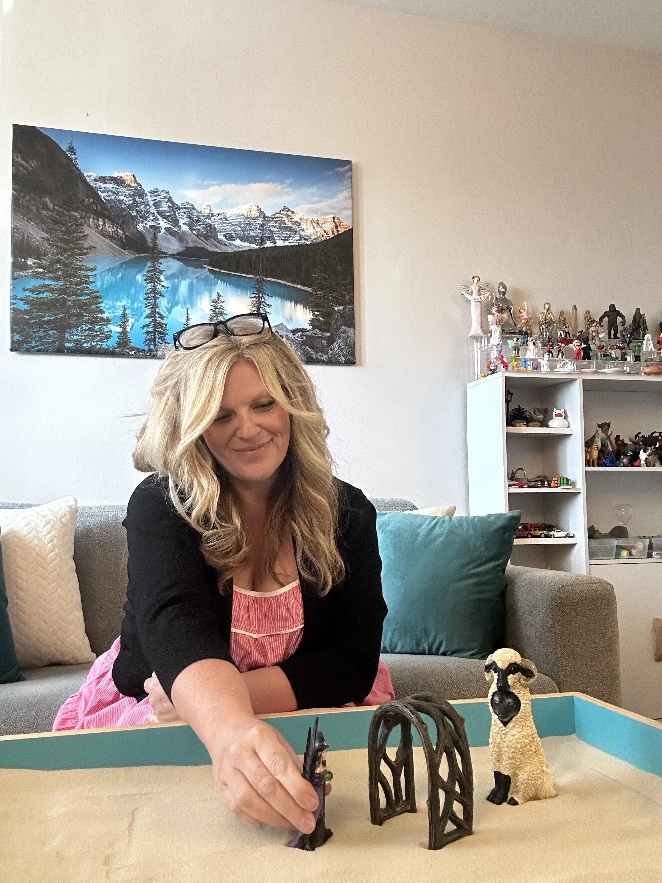 A woman with blonde hair and glasses on her head plays with toy figurines on a table in a living room, with a colorful sheep figure and a fantasy-themed toy scene. There is a couch with pillows and a bookshelf with figurines and collectibles in the background, and a large landscape photograph of mountains and a lake hanging on the wall.