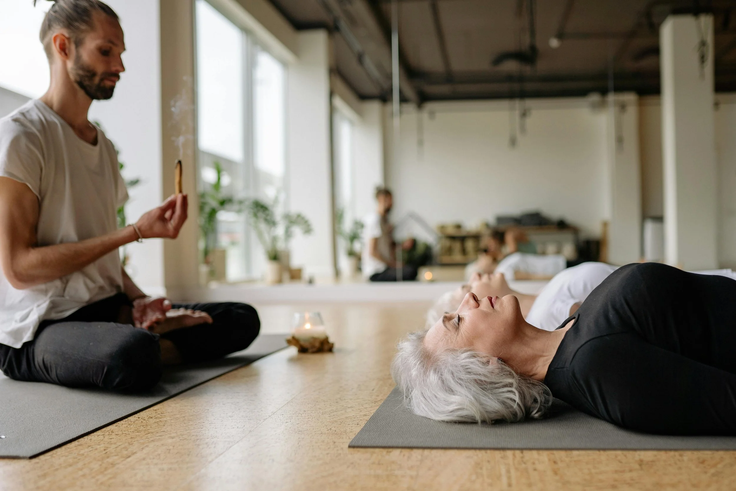 A woman lying on a yoga mat with eyes closed, in a yoga or meditation class, while an instructor sits nearby, holding a snack, in a bright, spacious room with natural light and plants.