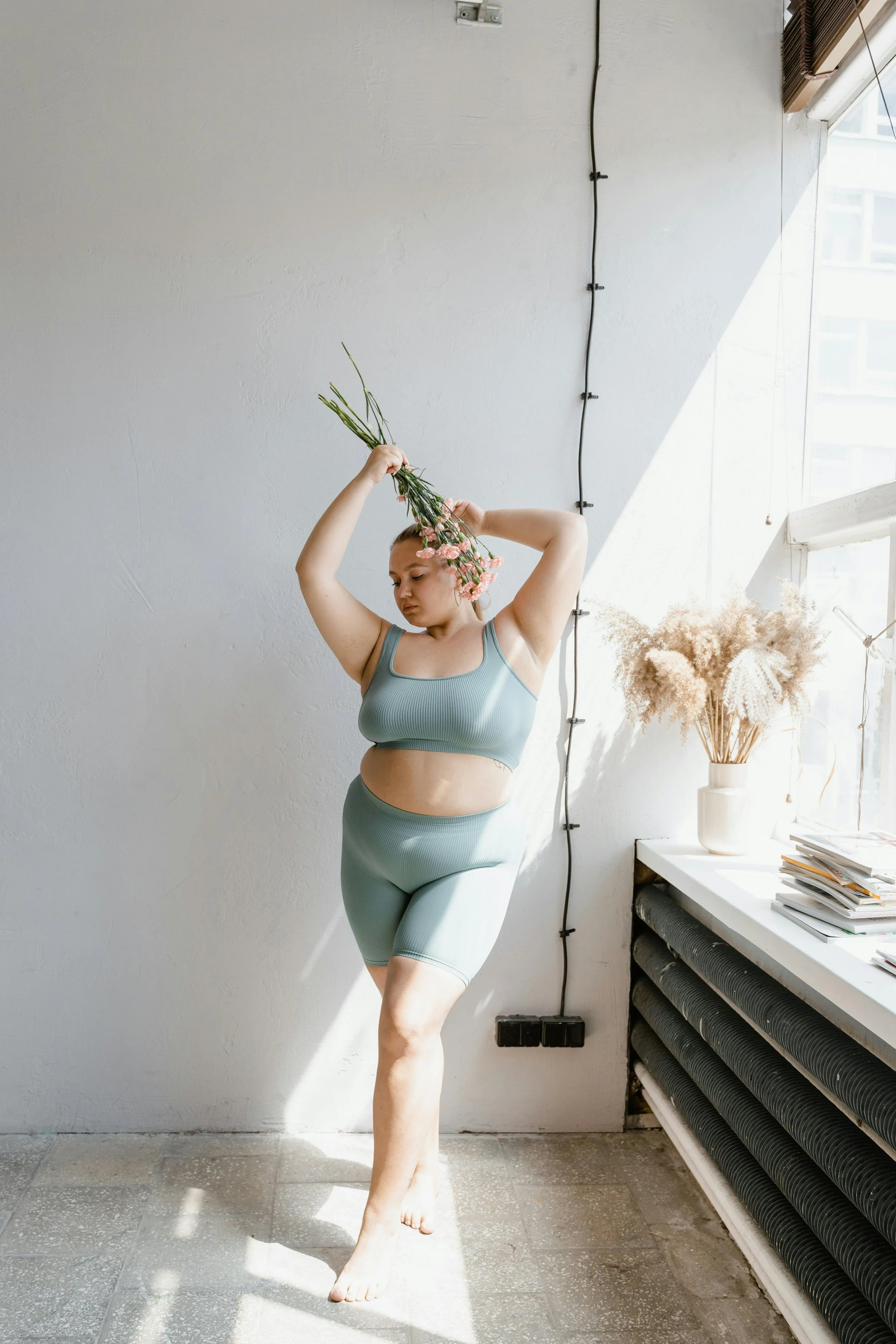 A woman in a light blue athletic outfit holding a bunch of pink flowers above her head, standing barefoot on a tiled floor in a room with natural light from a large window.