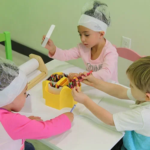 Three children wearing protective hair covers and masks are writing with markers at a table. One girl in a pink shirt is sitting, while another girl in a pink shirt with long sleeves and a white headband is standing and holding a marker. A boy in a white shirt and glasses is handing a marker to the girl.