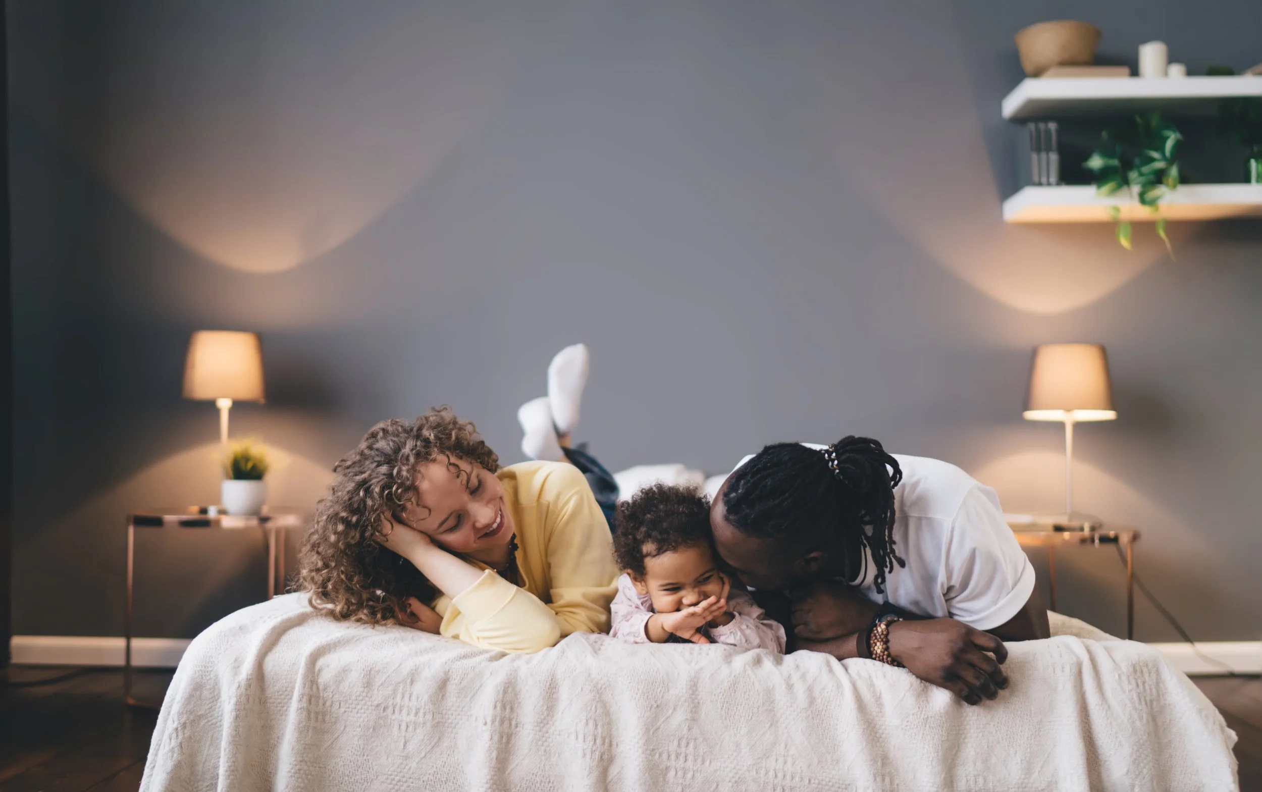 A family of three, with a woman, a man, and a young girl, lying on a bed and laughing together in a cozy bedroom.