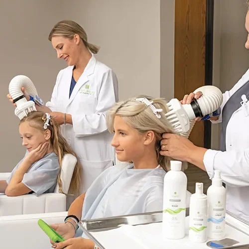 Two children getting their hair dried with hairdryers in a medical or salon setting, with a woman using a handheld device and another woman holding a large hairdryer, both wearing white coats.
