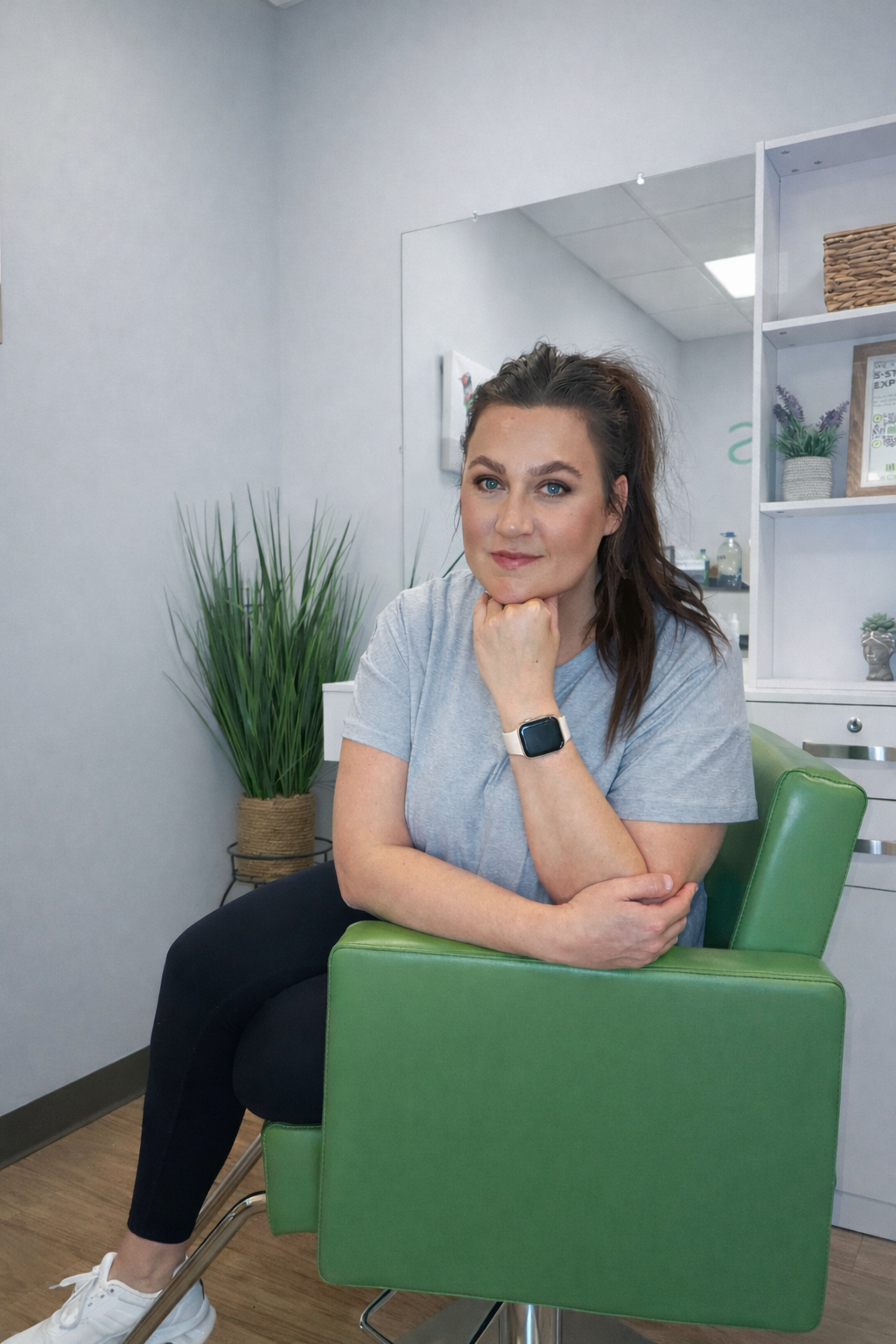 A woman with brown hair, wearing a gray t-shirt and a smartwatch, sitting on a green chair, resting her chin on her hand, looking at the camera, with a plant in the background inside a cozy indoor space.