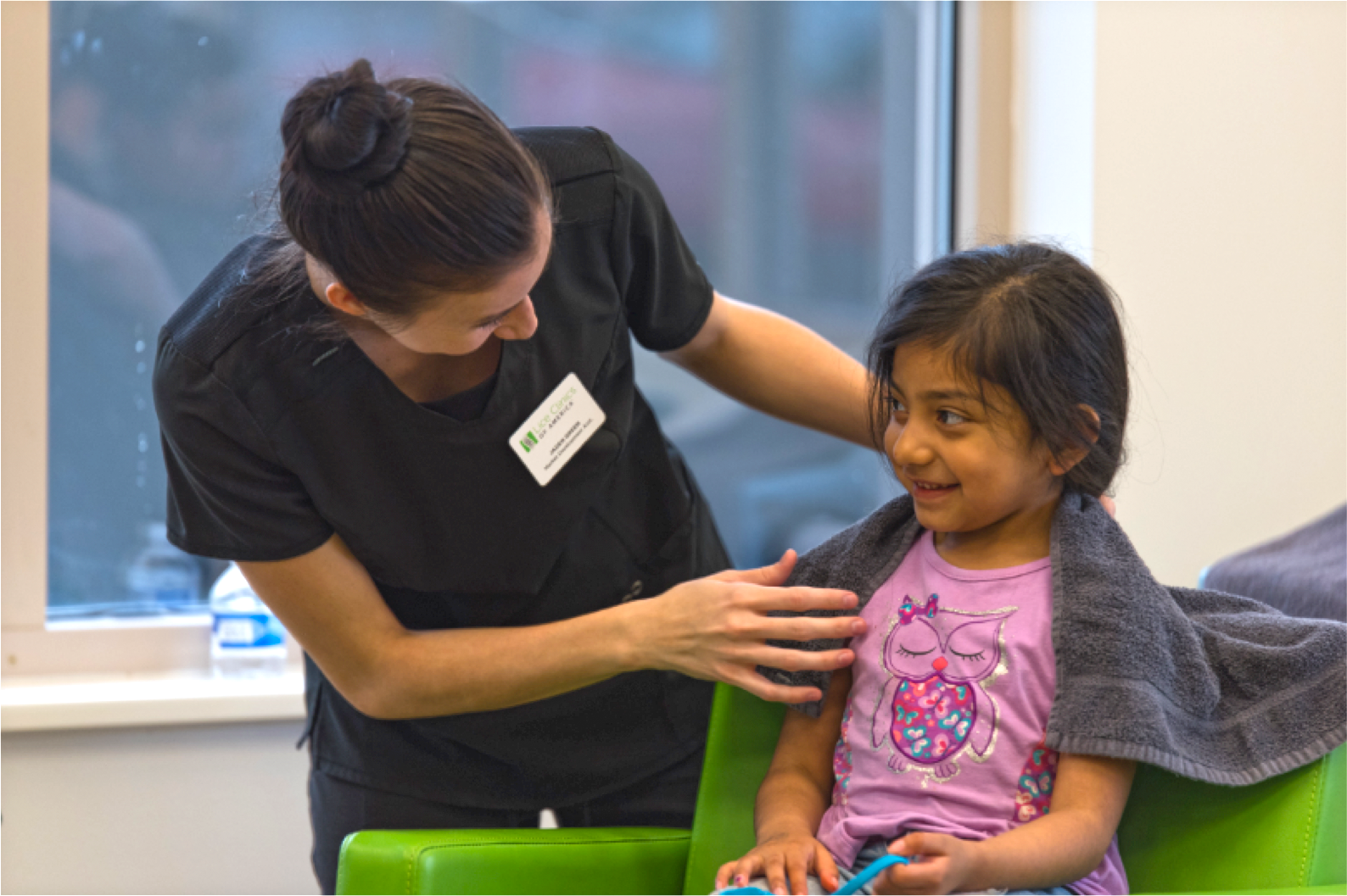 A young girl sitting in a green chair, smiling, with a gray towel over her shoulders, talking to a healthcare professional who is leaning over her, with a warm expression inside a medical office.