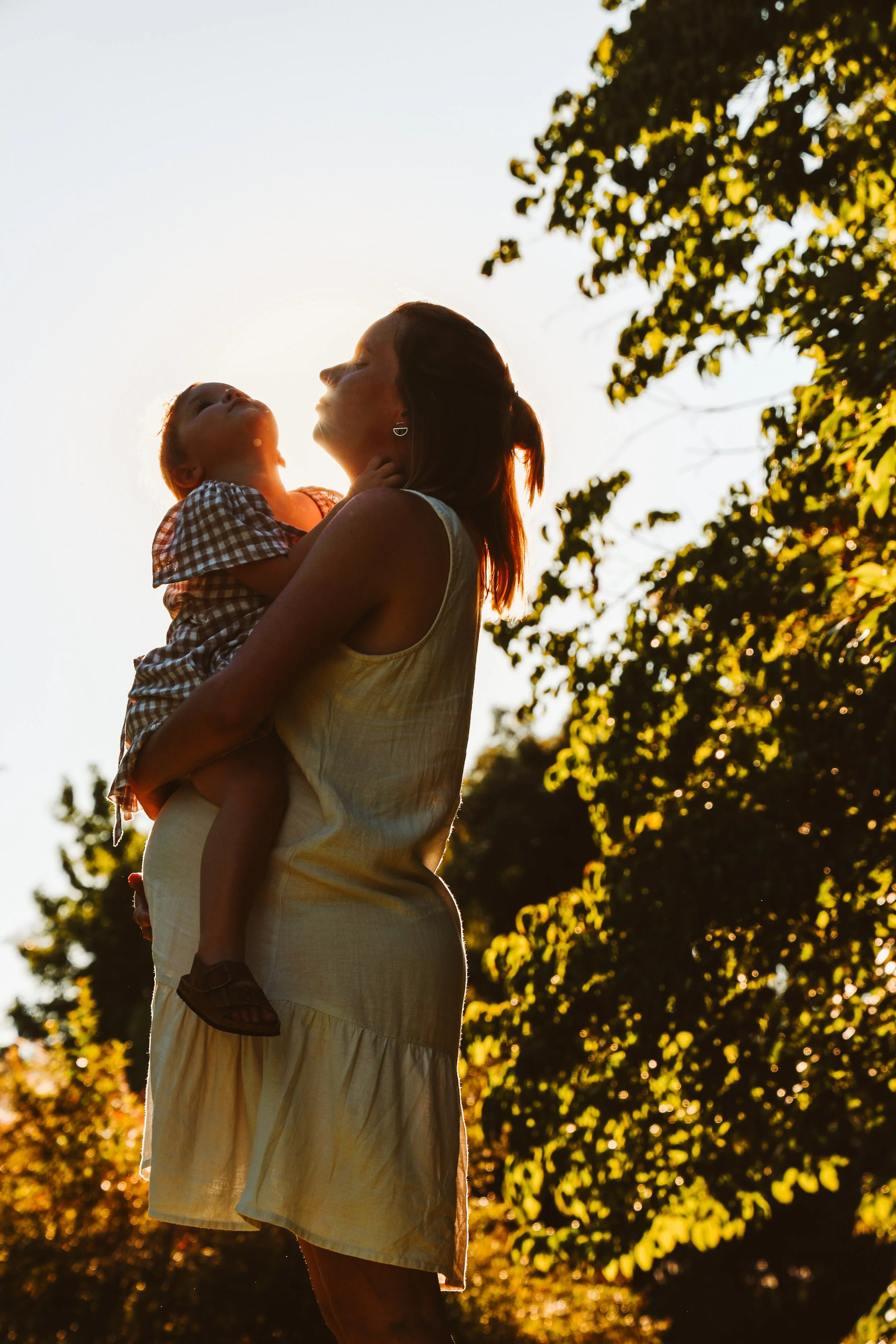 Mother lifting and smiling at her young child in warm sunset light beneath leafy trees.