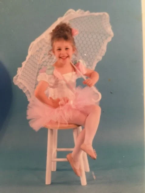 A young girl sitting on a small white chair, dressed in a pink tutu, holding a lace parasol, smiling at the camera against a solid light blue background.